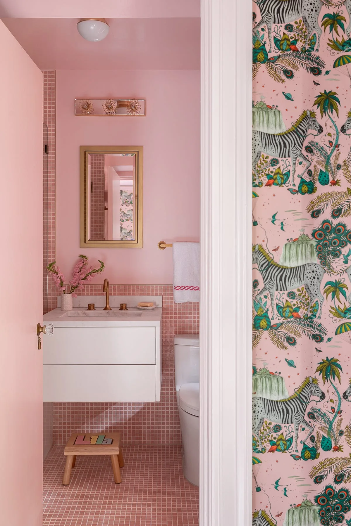 A pink powder room viewed through a doorway, showing pink mosaic tiles, a white floating vanity, a gold-framed mirror, and zebra wallpaper.