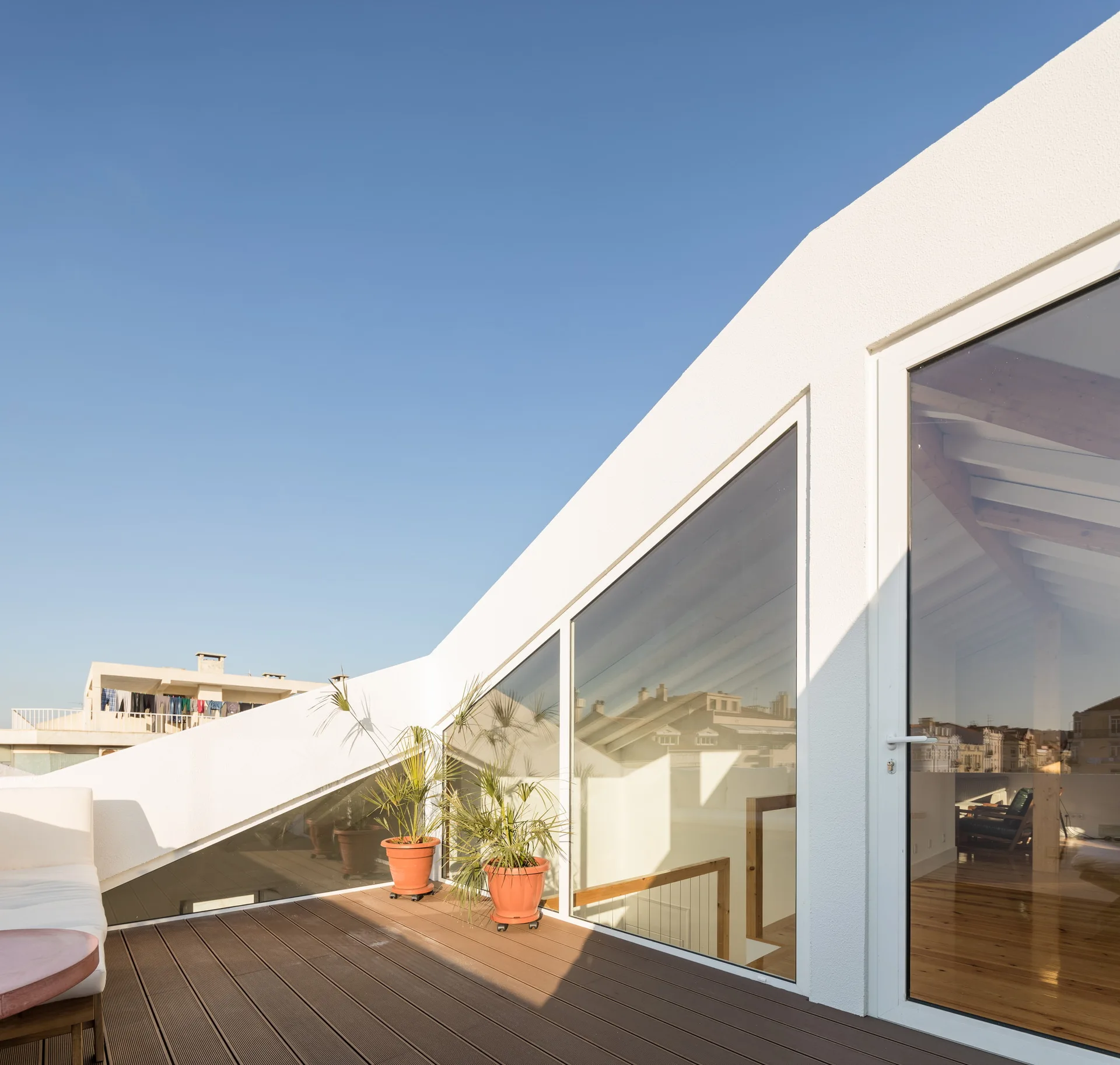 A sunny rooftop terrace features brown wood plank decking, smooth white stucco walls, large geometric windows, and potted green plants.