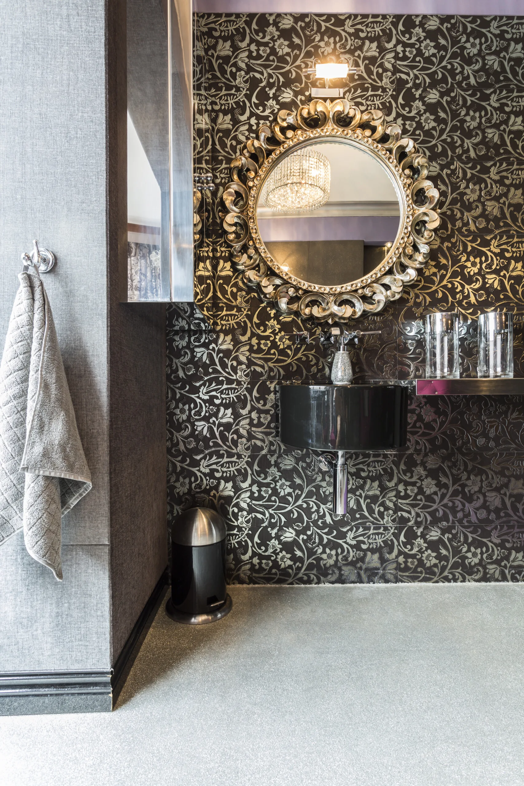 A powder room features a black wall-mounted sink set against intricate black and gold floral tiled walls beneath an ornate gold circular mirror.