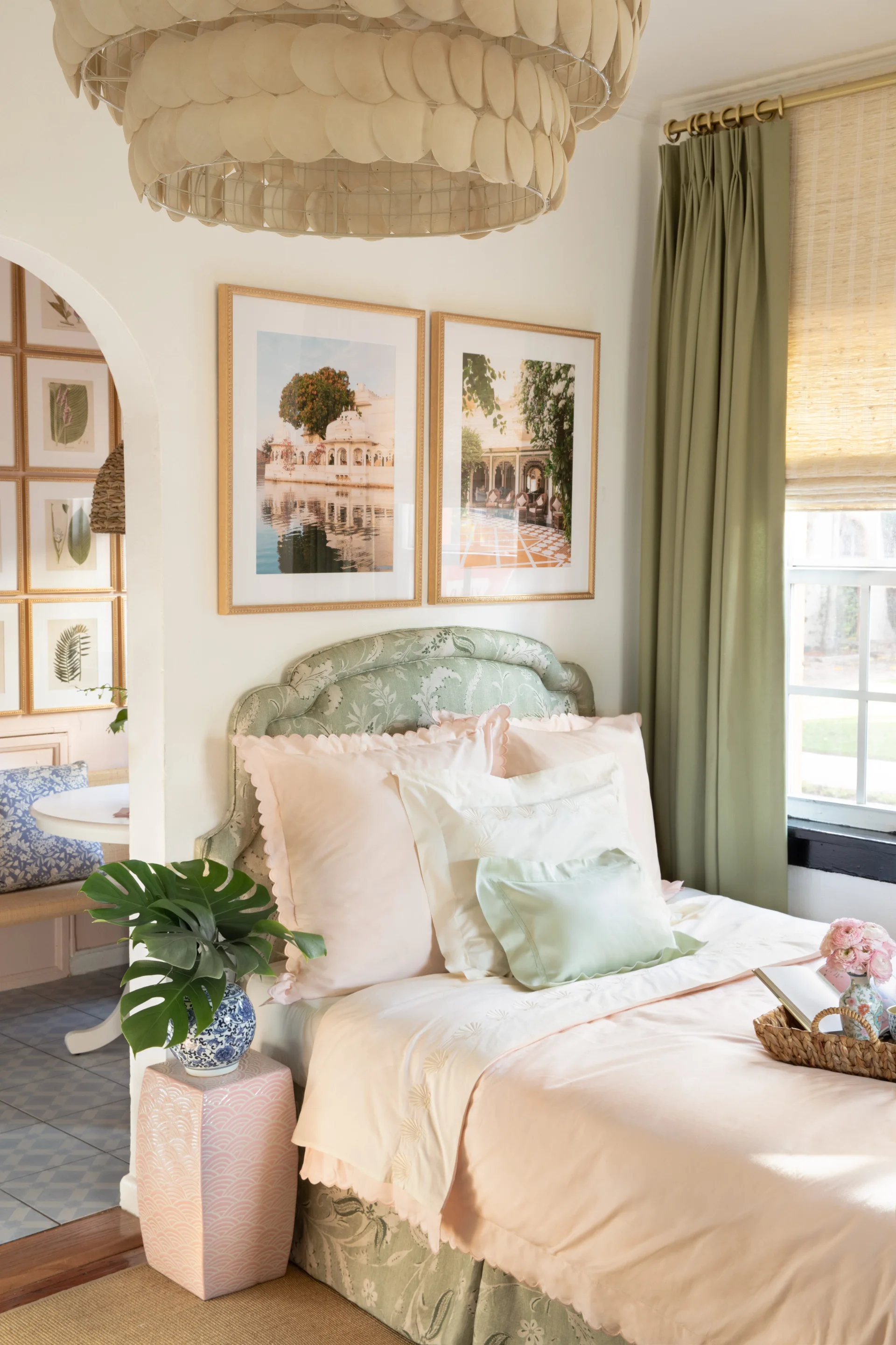 A bright bedroom features a green floral upholstered headboard, pink bedding, a pink garden stool nightstand, and sage green curtains.