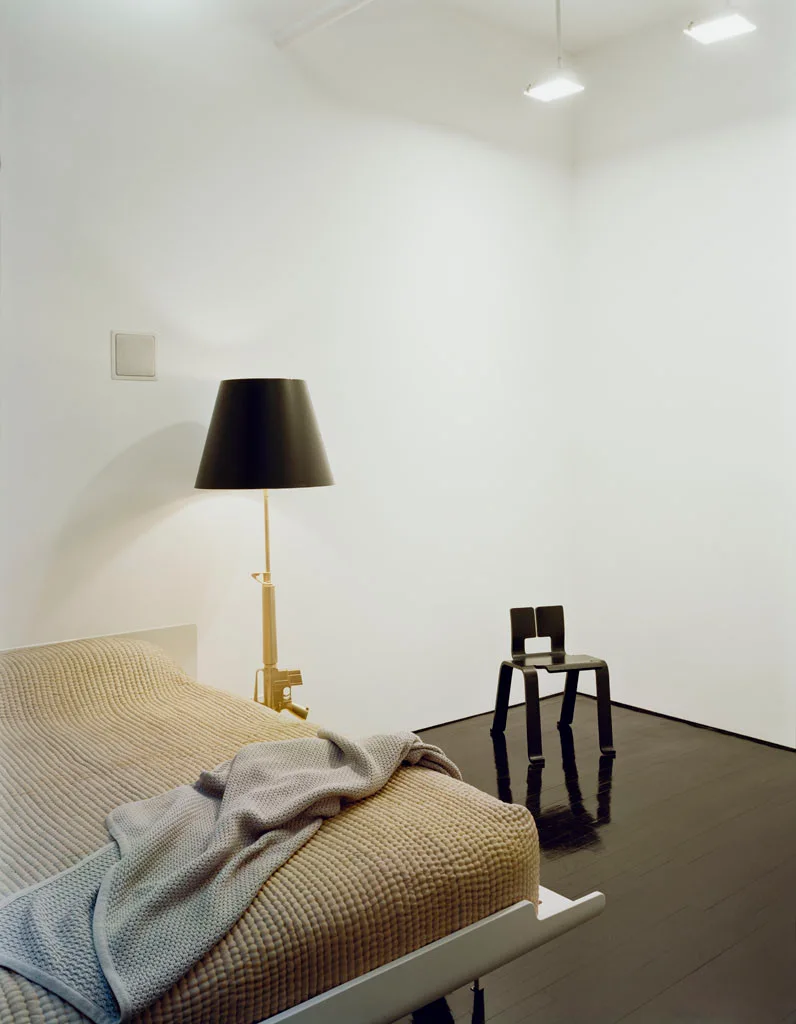 A minimalist bedroom featuring a bed with textured beige bedding, a gold rifle-base floor lamp, and a black chair on glossy black flooring.