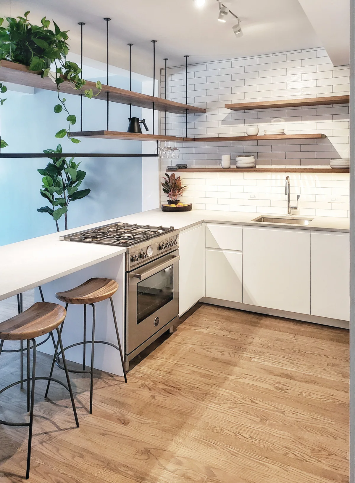 A modern kitchen featuring a white island with a built-in stove, wooden bar stools, floating shelves, and a white subway tile wall.
