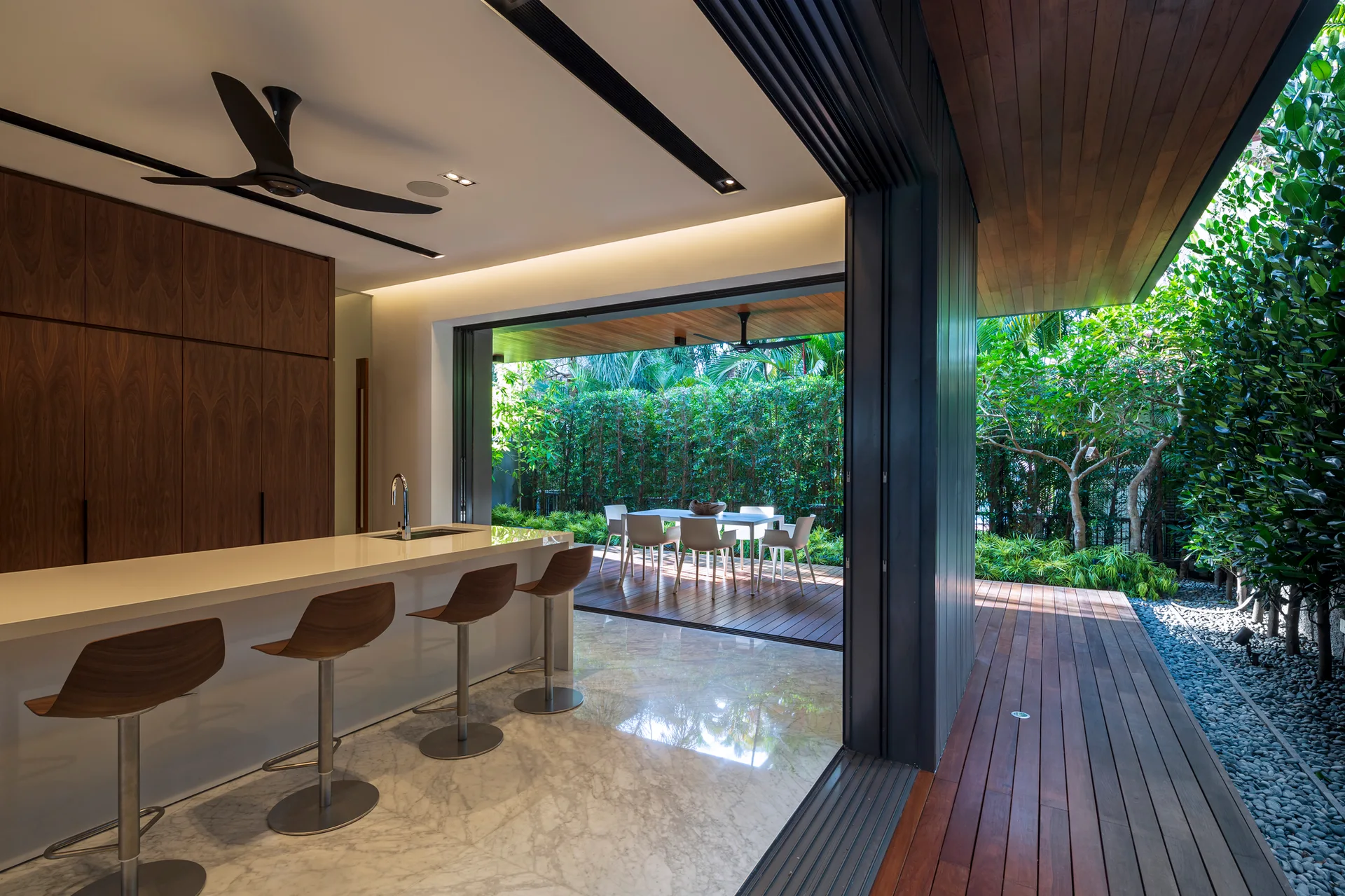 A modern kitchen with marble floors and wood cabinetry opens completely onto a wood patio deck with an outdoor dining table.