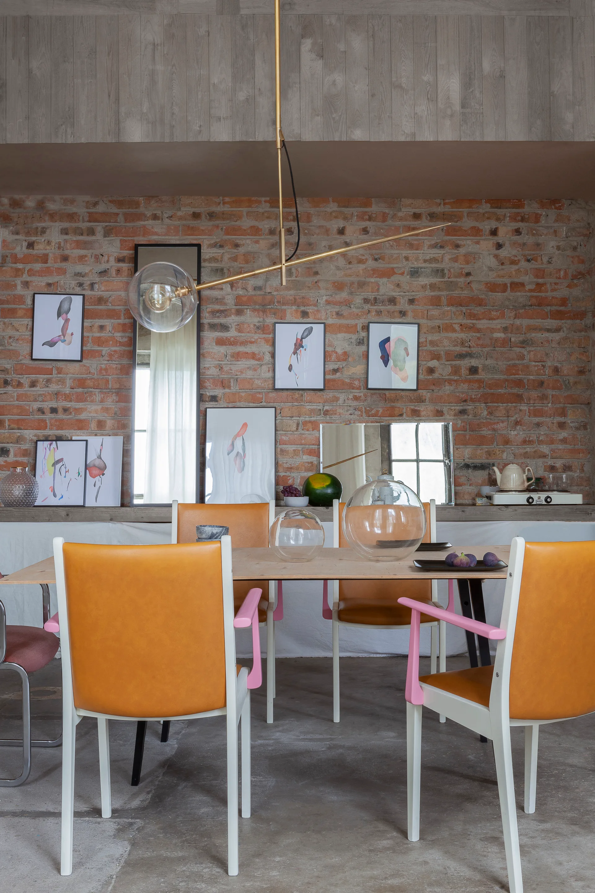 A dining room featuring a light wood table, orange and white chairs with pink armrests, and an exposed brick wall decorated with small framed artworks.