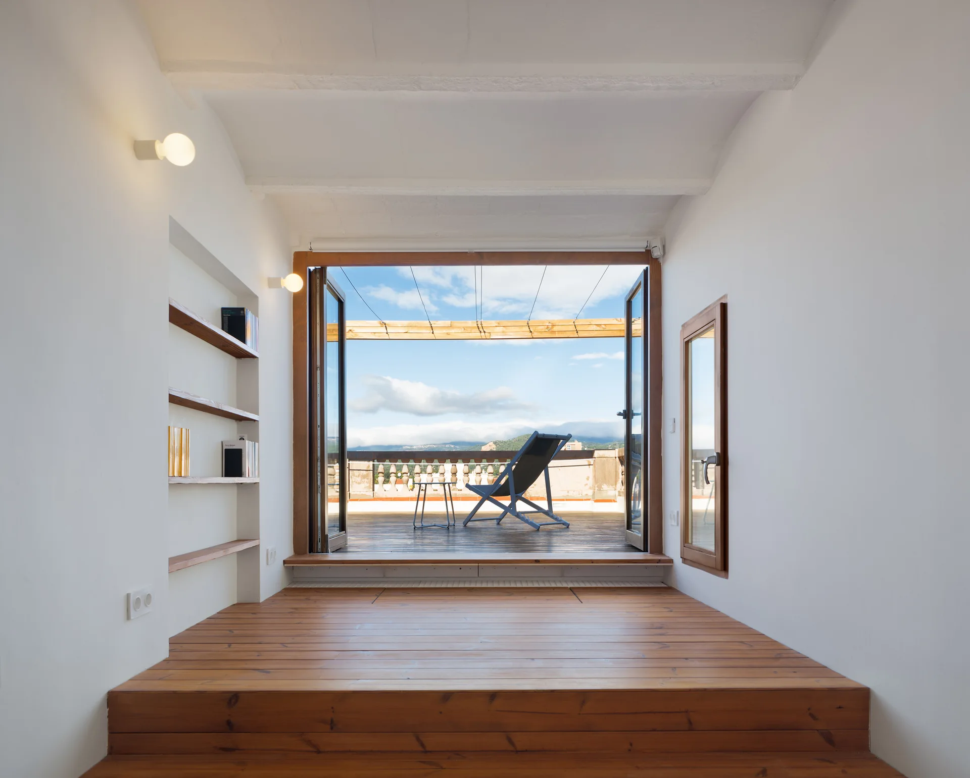 A minimalist room features a raised wooden floor transitioning through open glass doors to a sunny outdoor terrace with a deck chair.