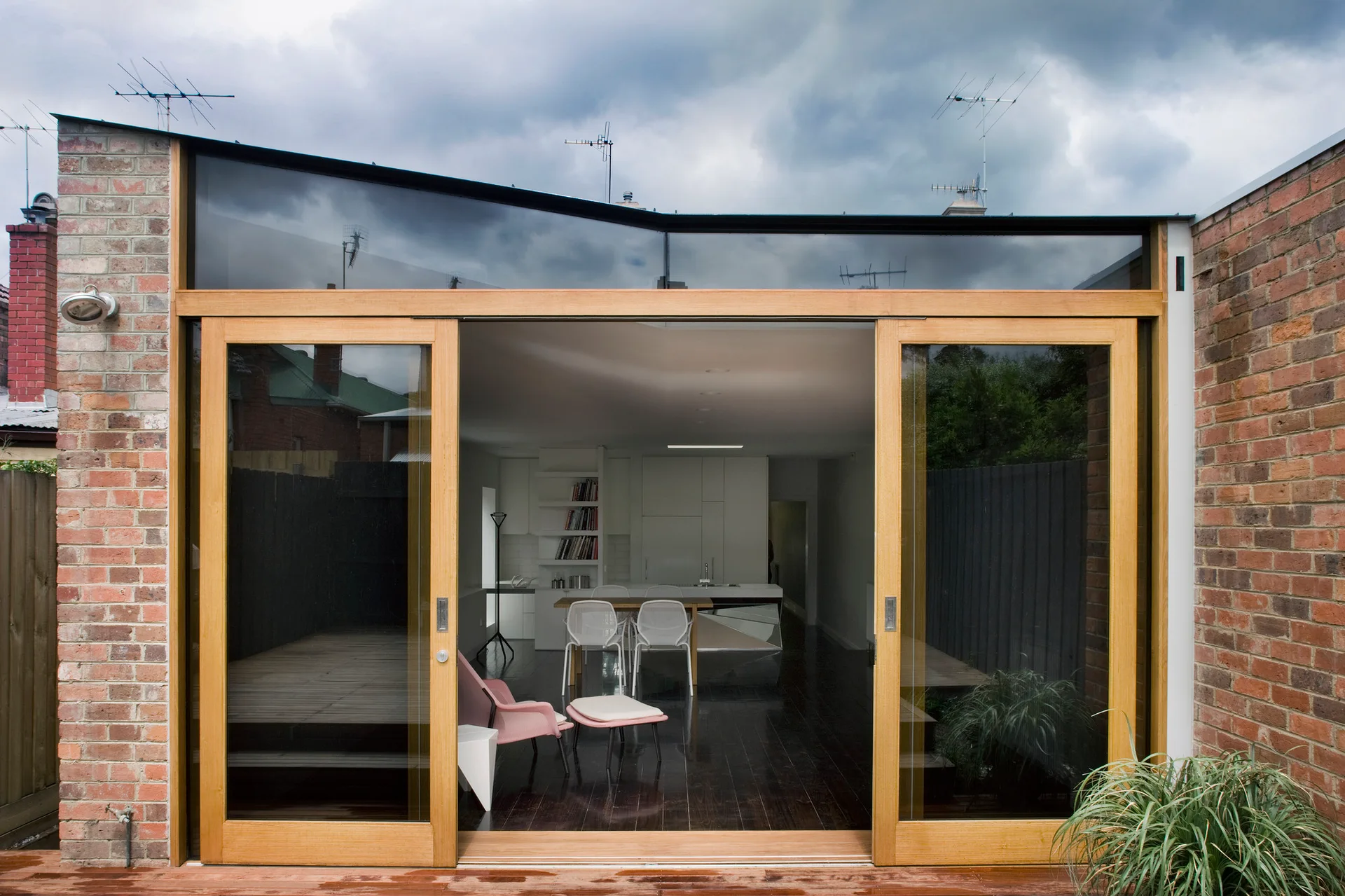 Large timber-framed sliding glass doors open from a brick patio into a minimalist white kitchen and dining area with dark wood floors.