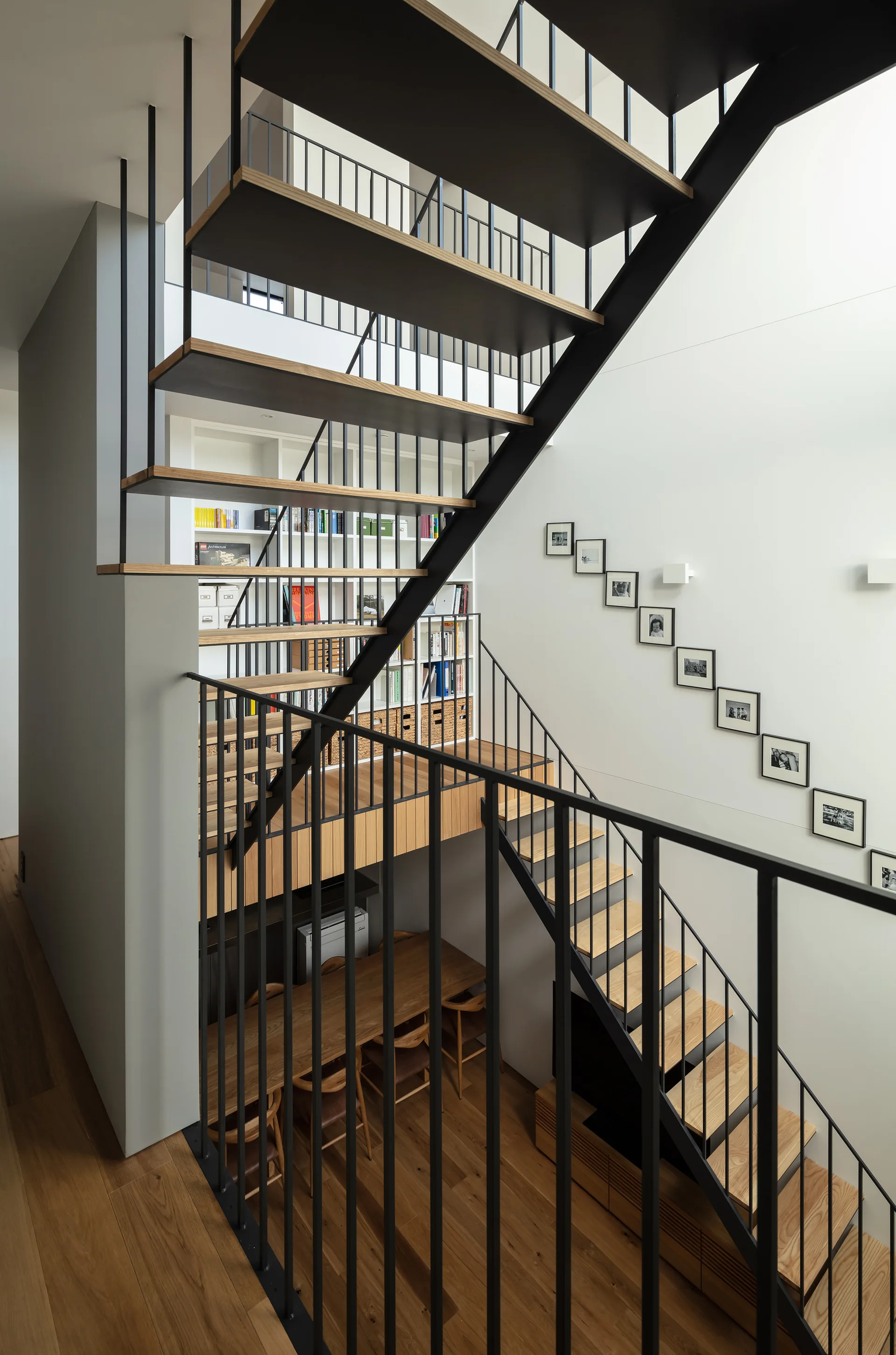 A modern multi-level staircase with black metal railings and wood treads rises above a dining table and built-in bookshelf.