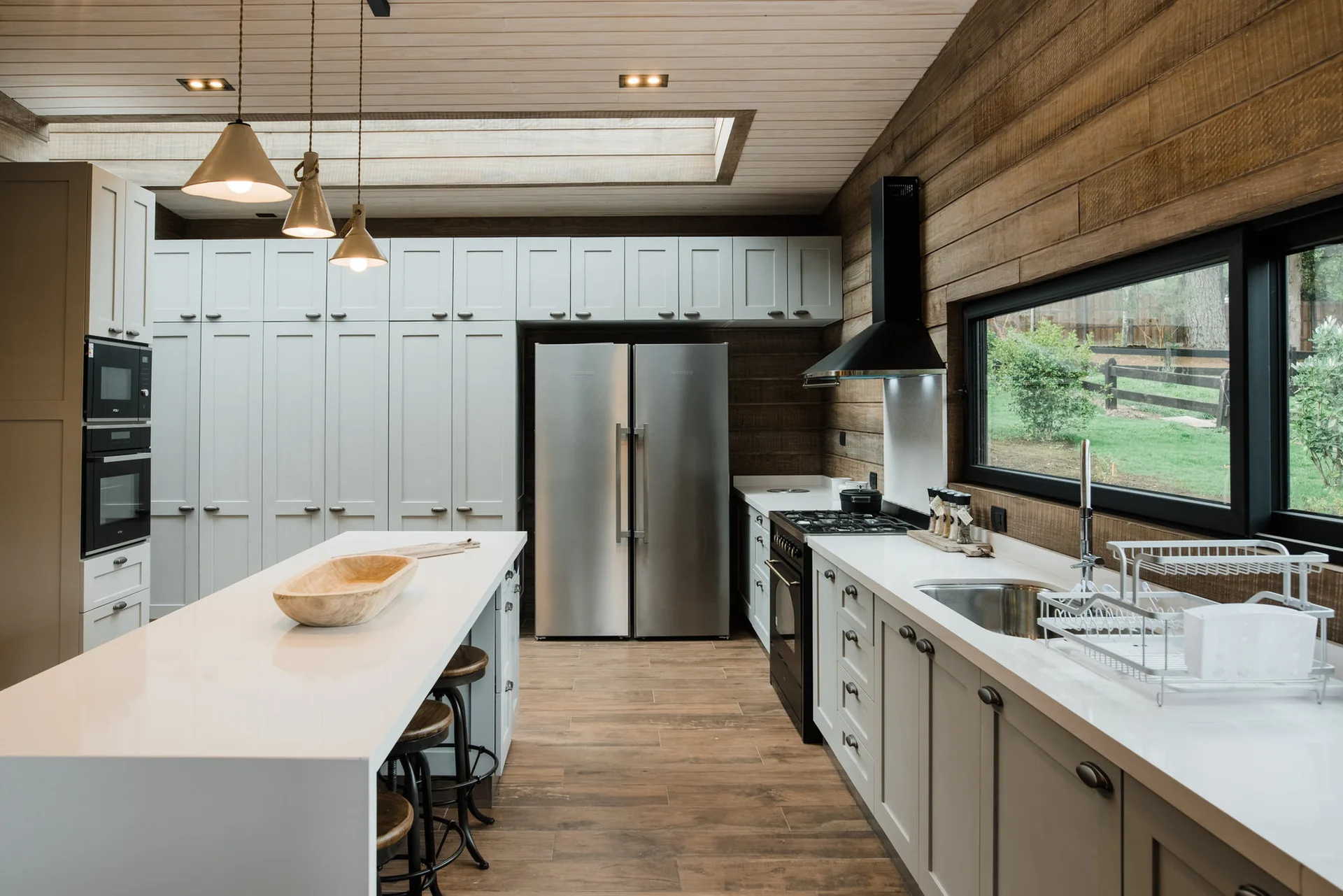 A modern farmhouse kitchen featuring light gray shaker cabinetry, a white island, stainless steel refrigerator, and horizontal wood-paneled walls.