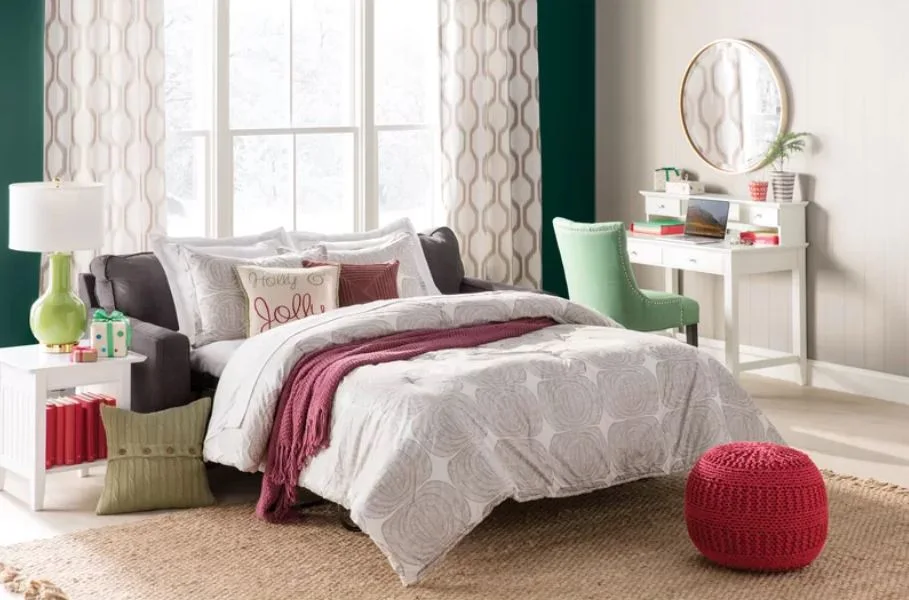 A dark gray sleeper sofa with red and white bedding sits beside a small white writing desk and a green accent chair.