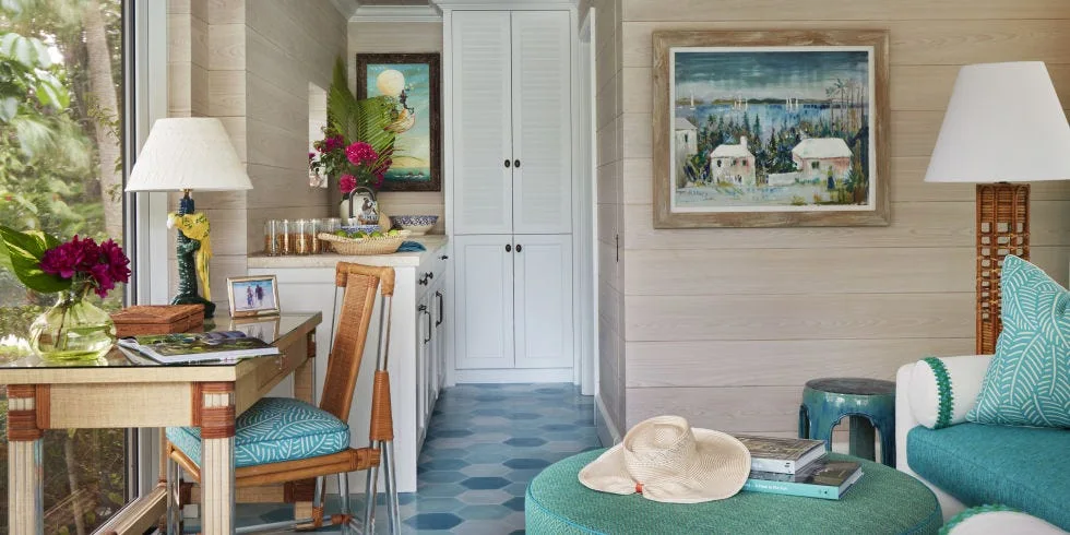 A coastal living room features blue hexagonal tile flooring, light wood paneled walls, a desk with a wicker chair, and a teal sofa.