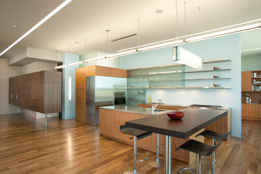 A modern kitchen features light wood cabinetry, integrated stainless steel appliances, and a dual-level island with dark bar stools on hardwood floors.