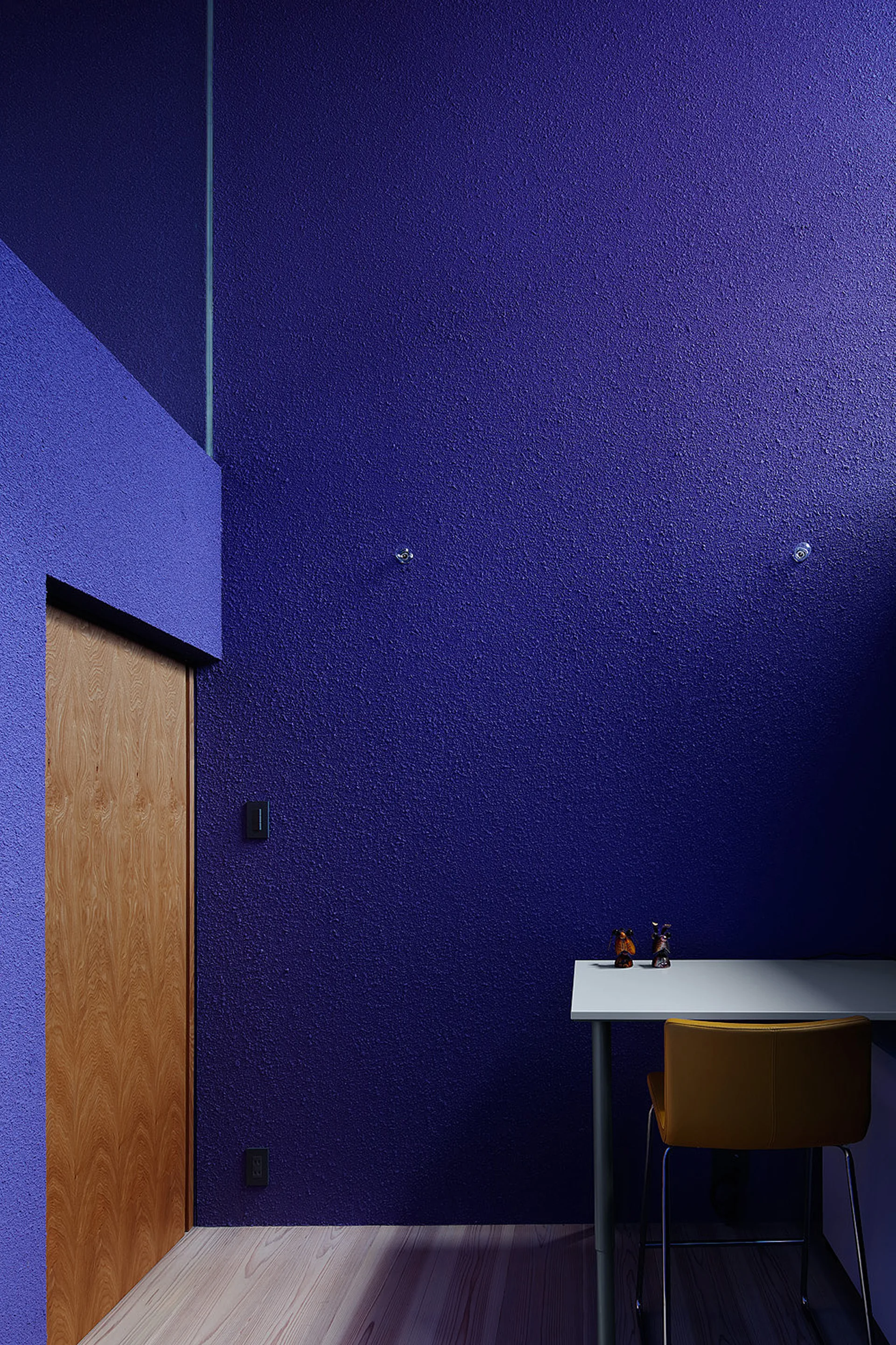 A minimalist workspace features a deep blue textured wall, a natural wood grain door, and a white desk paired with a yellow chair.