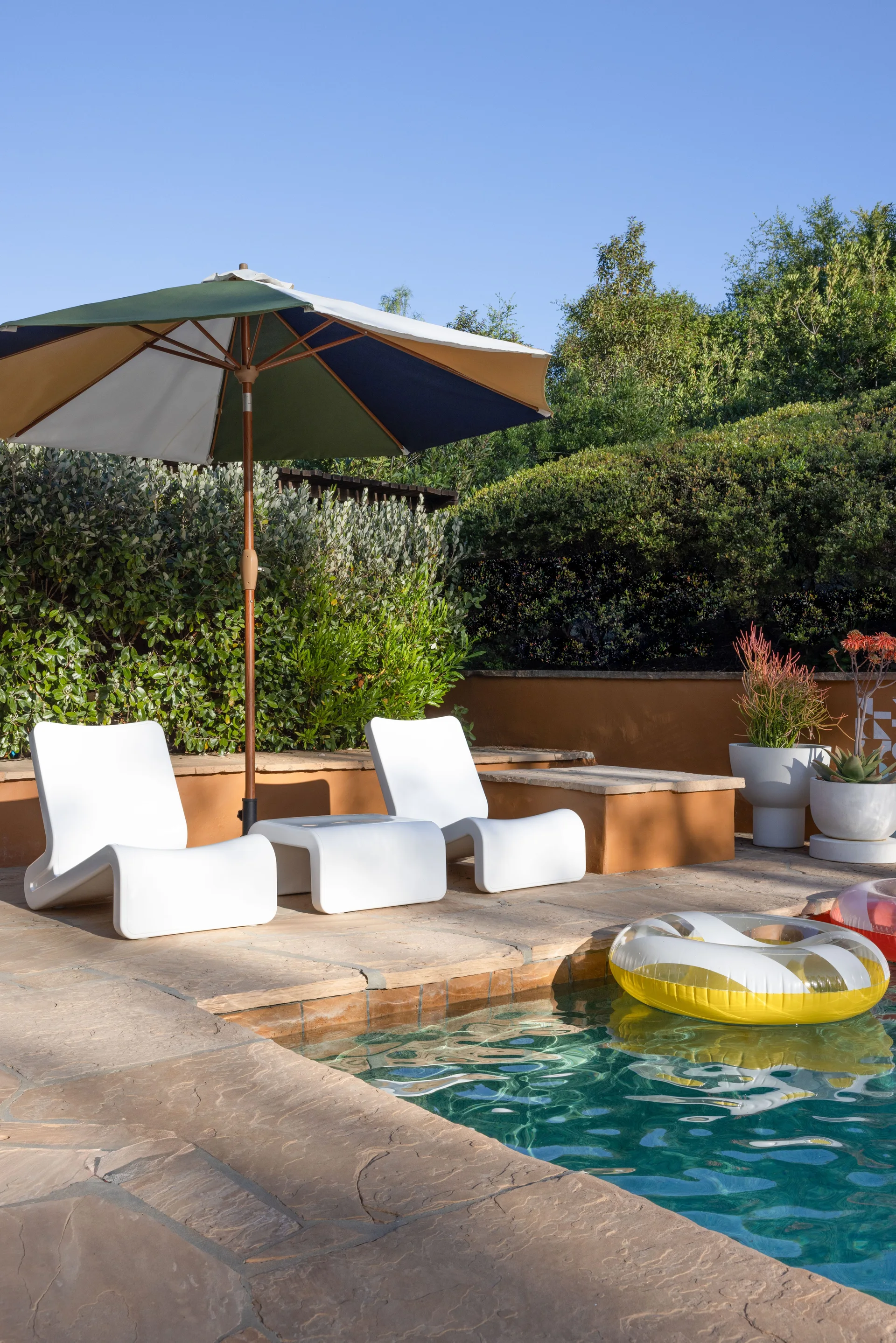 Two curved white lounge chairs and a multi-colored patio umbrella sit on a stone pool deck next to a swimming pool with lush green landscaping behind.