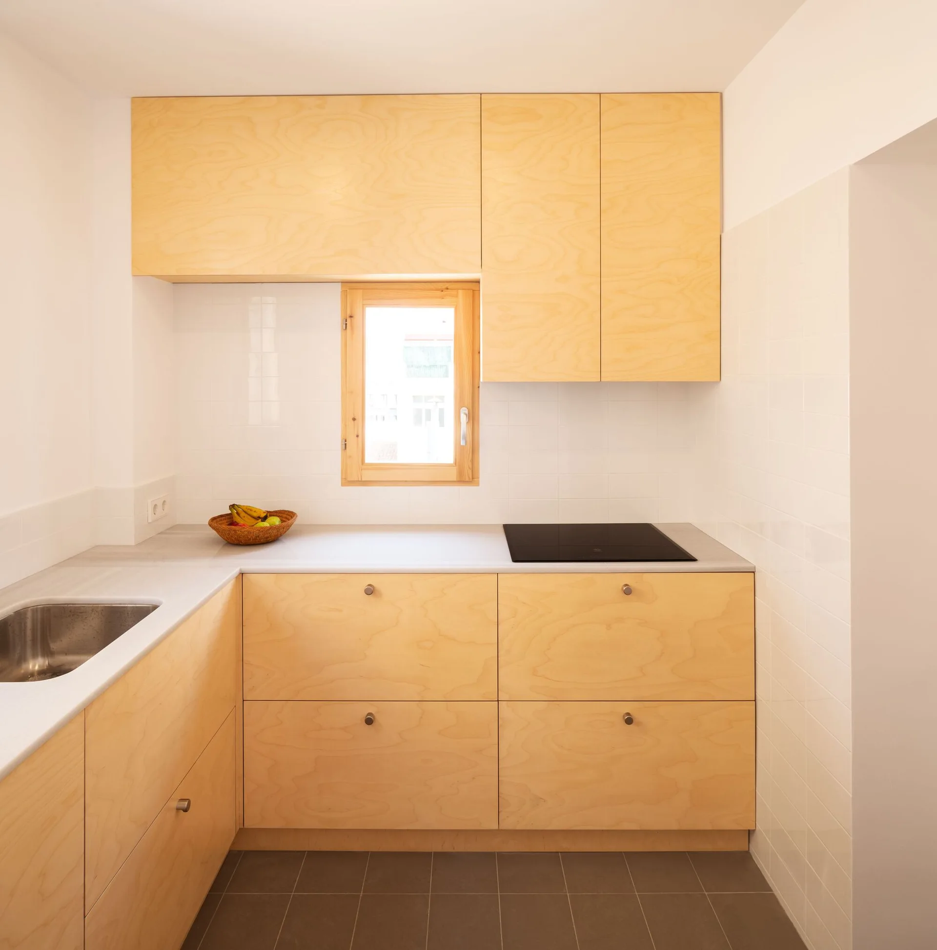 A minimalist kitchen features flat-front plywood cabinetry, a small wood-framed window, and a glossy white square tiled backsplash.