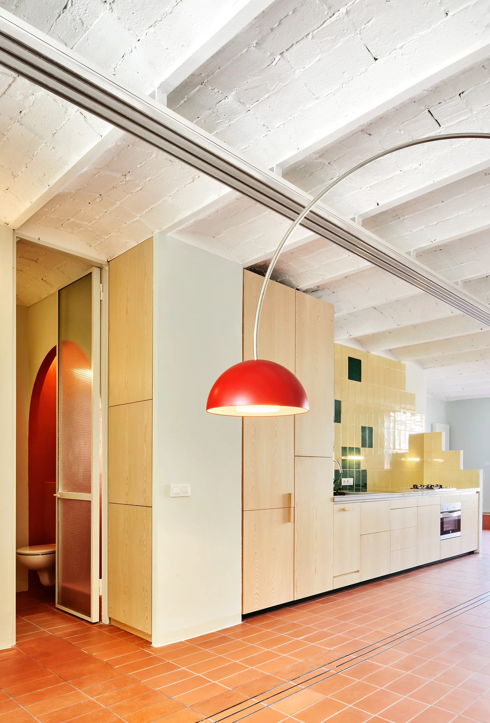 A modern kitchen features light wood cabinets, square terracotta floor tiles, a yellow tiled backsplash, and a bright red dome pendant lamp hanging from above.