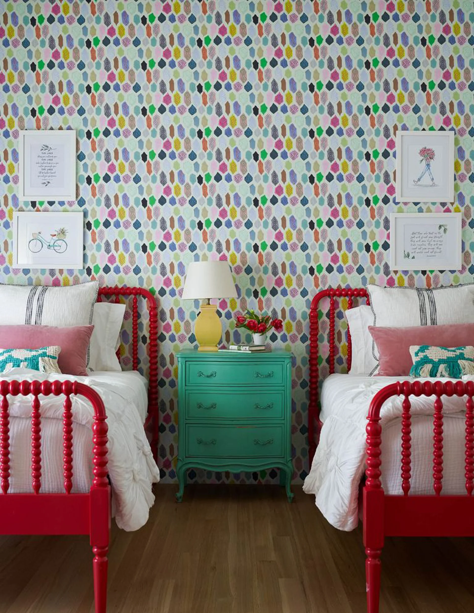 Two red wooden spool beds with white linens flank a teal nightstand in front of colorful geometric wallpaper.