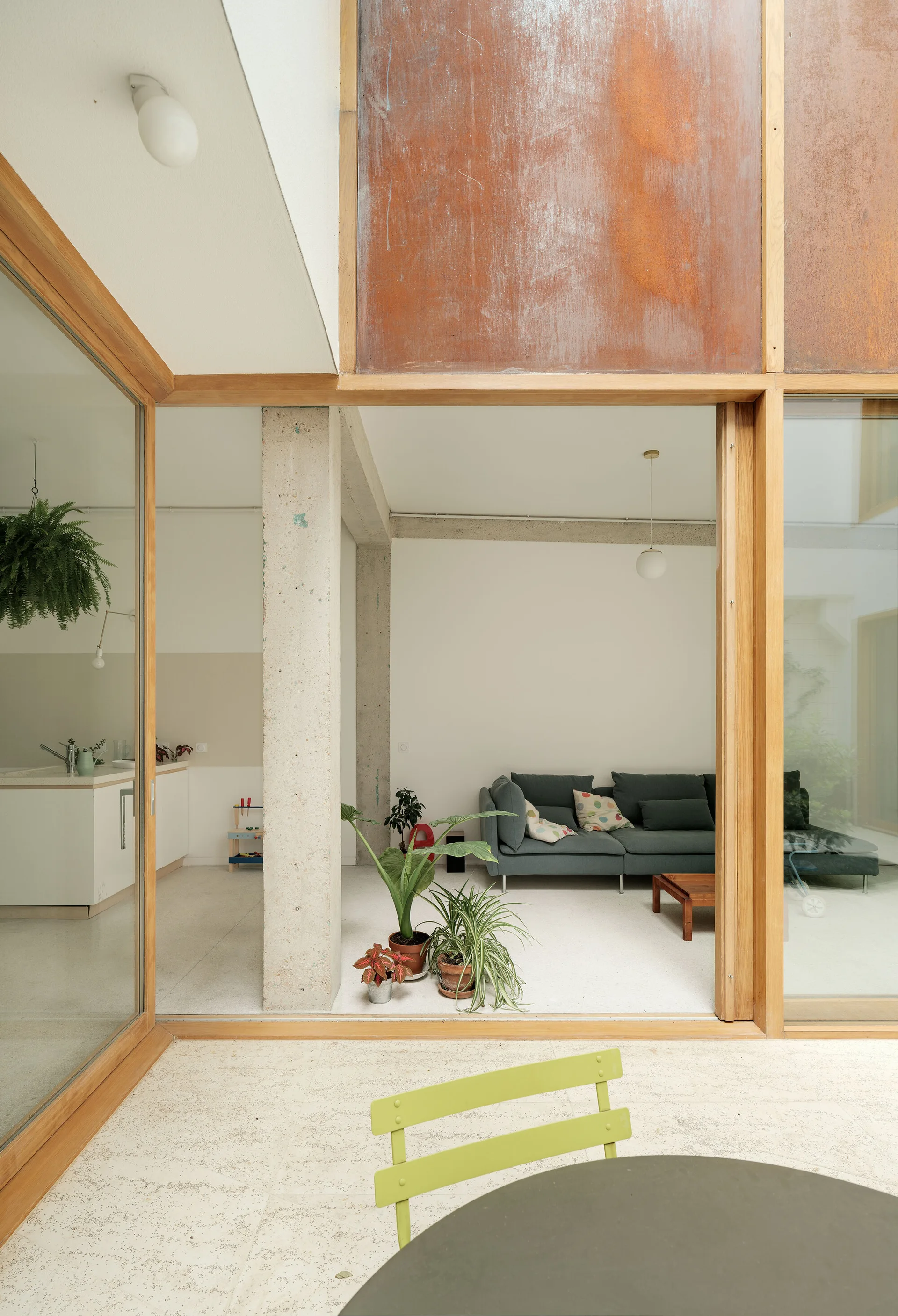 A modern living room with a grey sofa is seen through large wood-framed glass sliding doors next to an exposed concrete pillar and weathered rust panel.