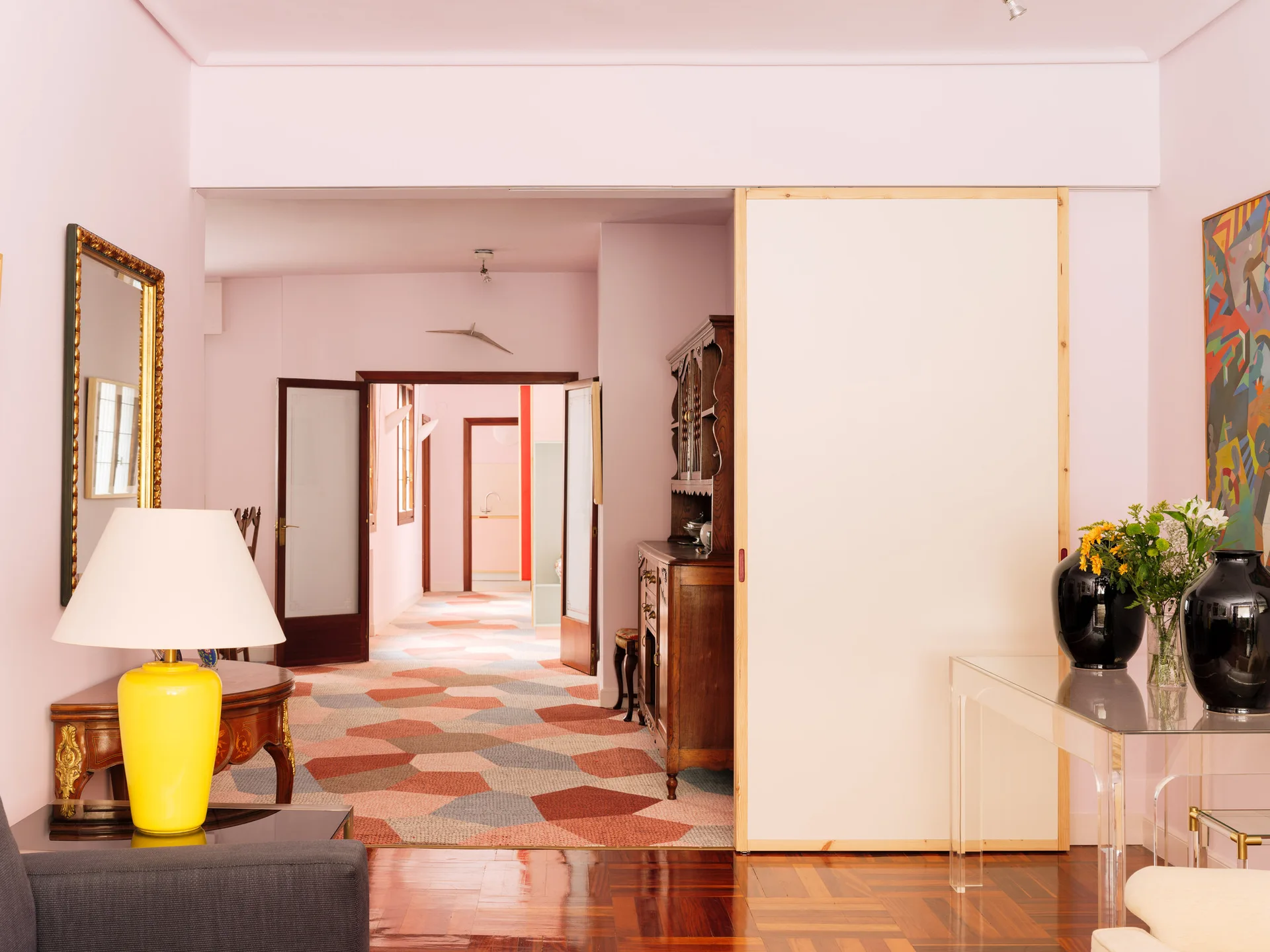 An interior space with pink walls and a wooden floor transitions into a hallway with multi-colored geometric tiles, featuring a bright yellow table lamp.