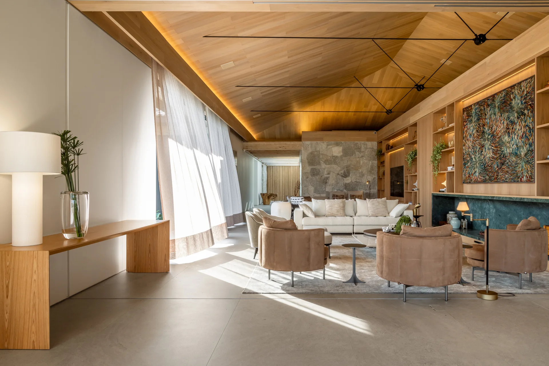 A spacious living room features a vaulted timber ceiling, tan armchairs, a white sofa, and a natural stone accent wall in the background.