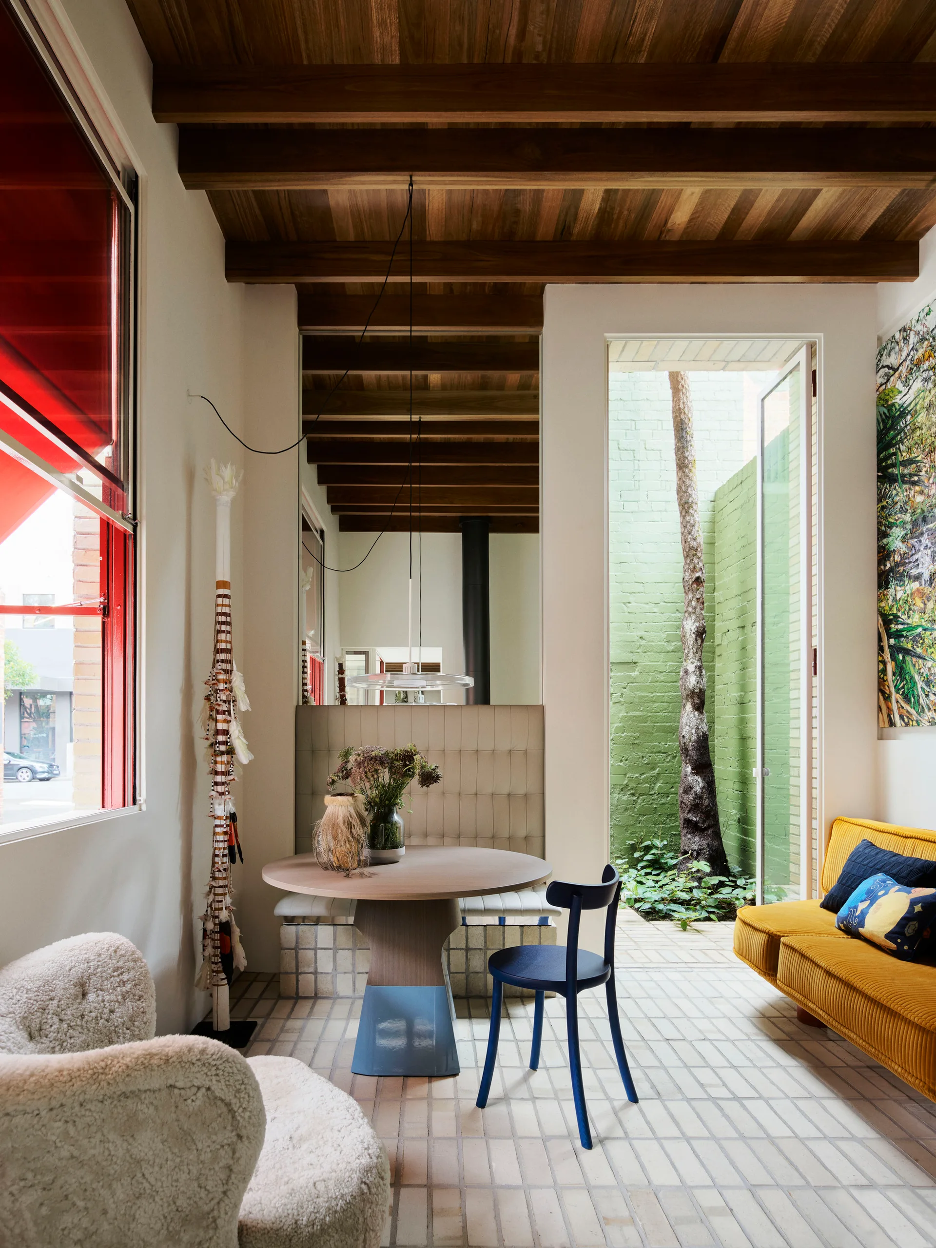 A modern living and dining room with a round wooden table, a yellow corduroy sofa, exposed ceiling beams, and a view of a small green courtyard.