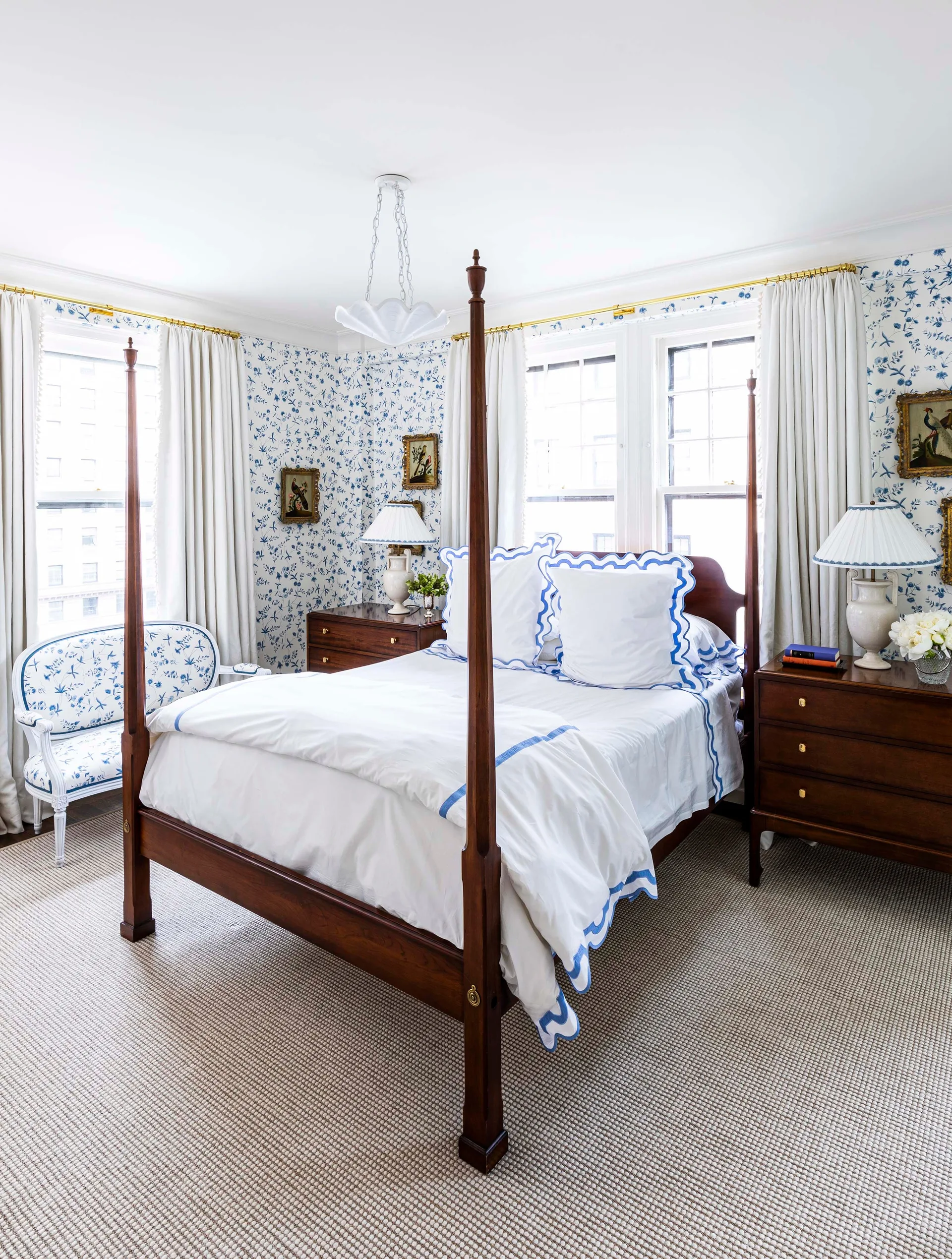 A traditional bedroom features blue and white floral wallpaper, a dark wood four-poster bed, and matching wooden nightstands under white drapes.