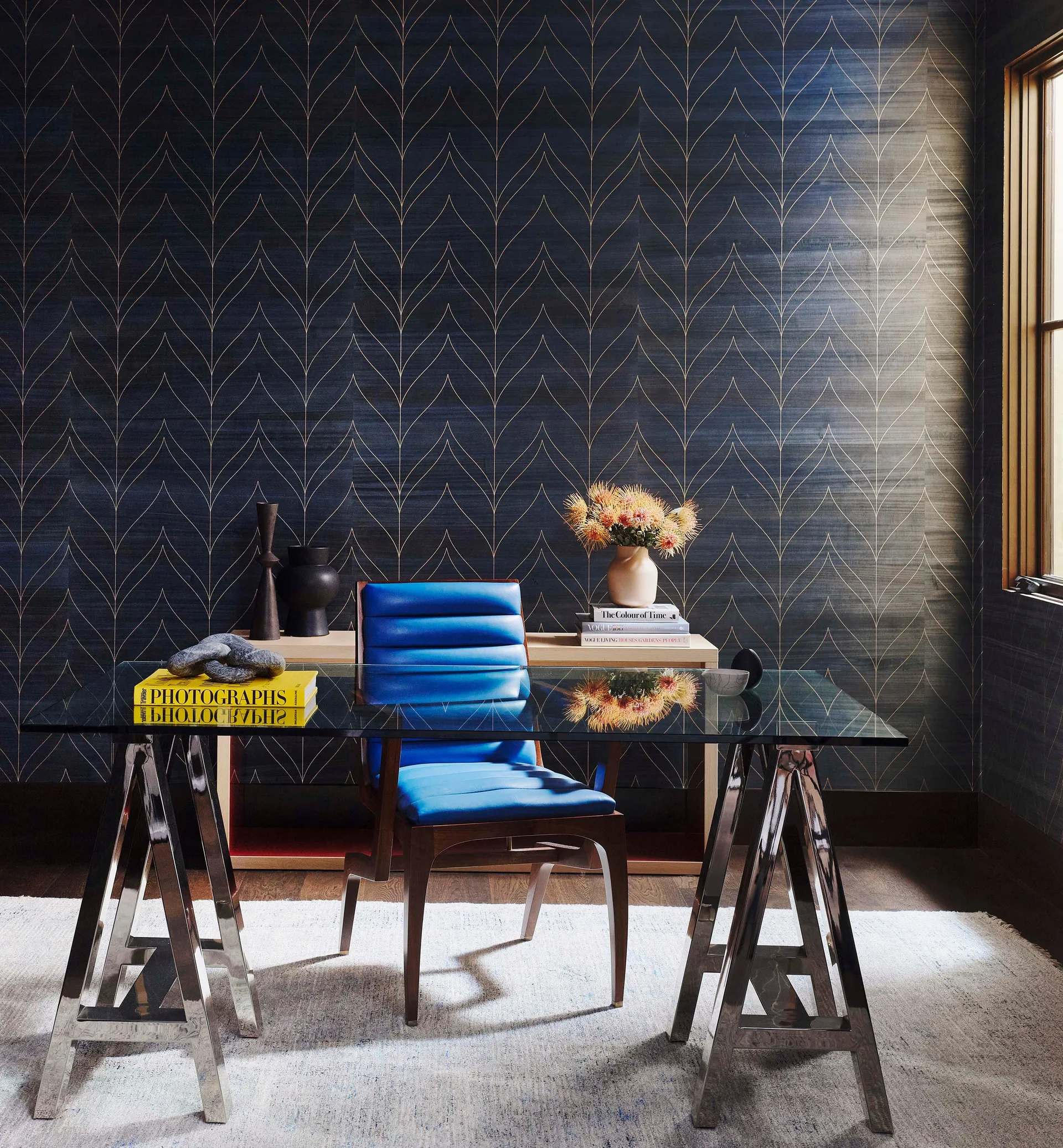 A home office features a glass-top trestle desk and a bright blue chair set against a navy blue wallpaper wall with a gold geometric pattern.