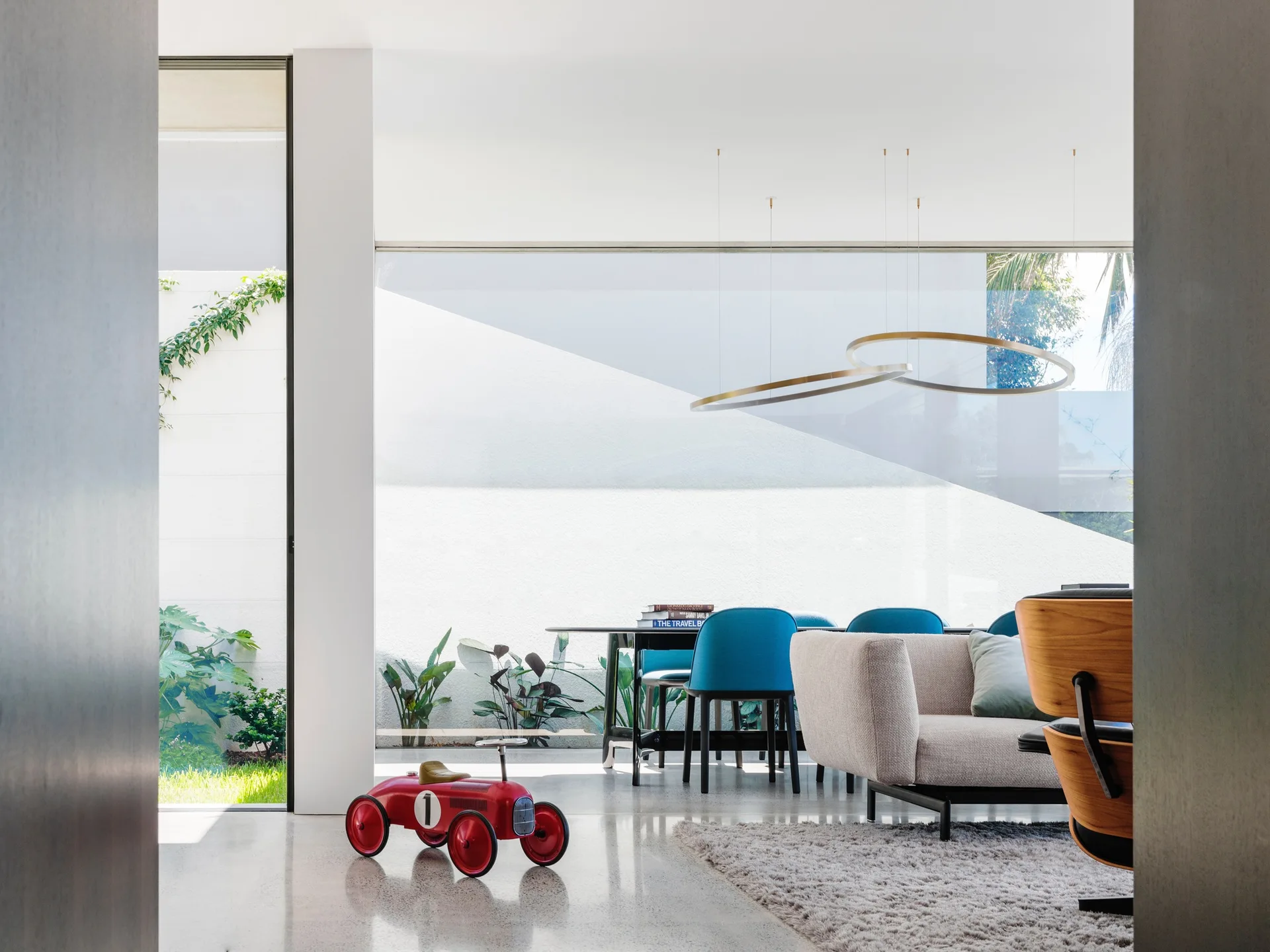 A modern open-plan living and dining area features teal chairs, a grey sofa, a gold ring pendant light, and a large window looking onto a courtyard.