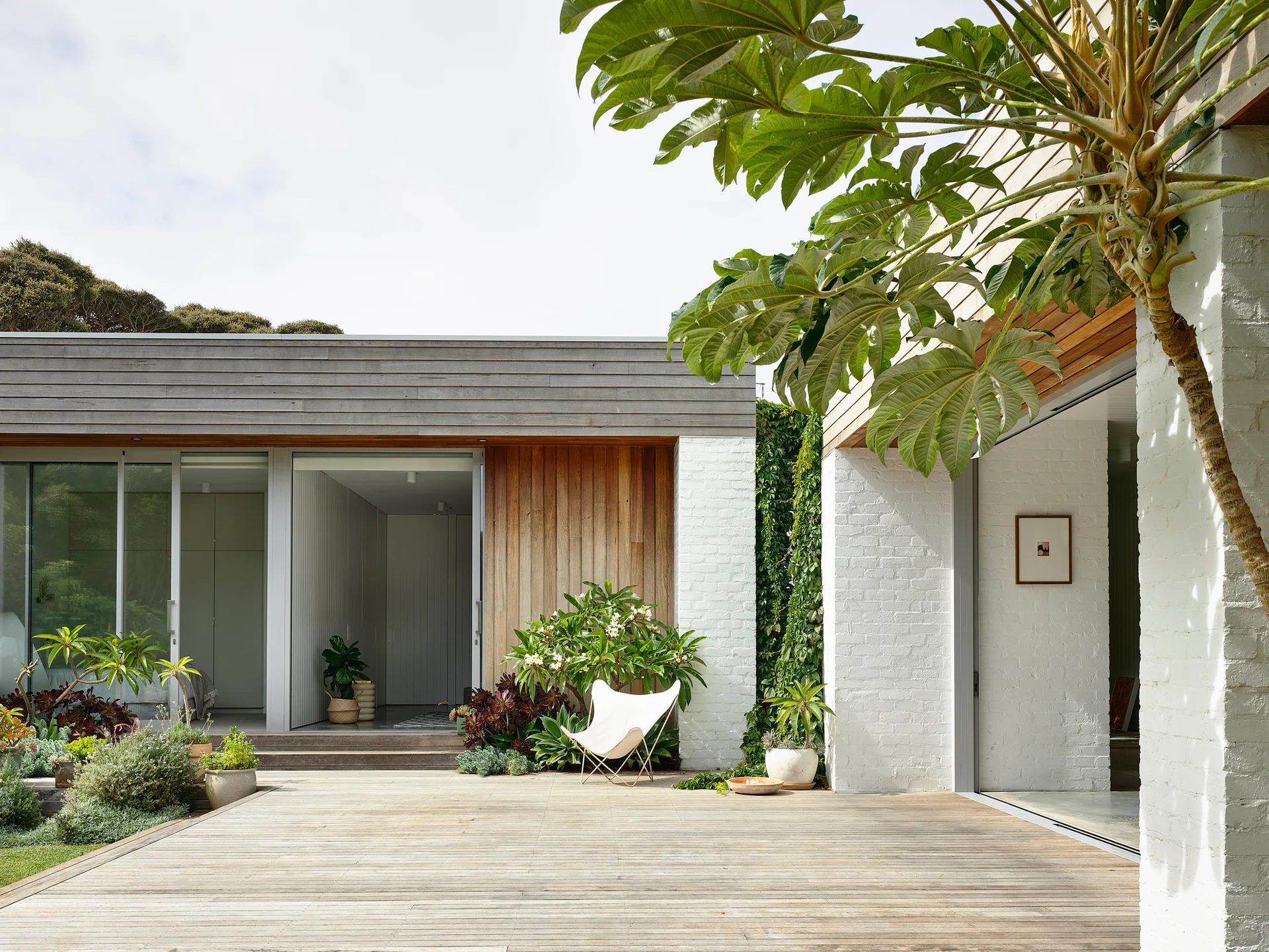 A modern outdoor patio features a wide wooden deck, white painted brick walls, natural wood siding, and a white canvas butterfly chair.