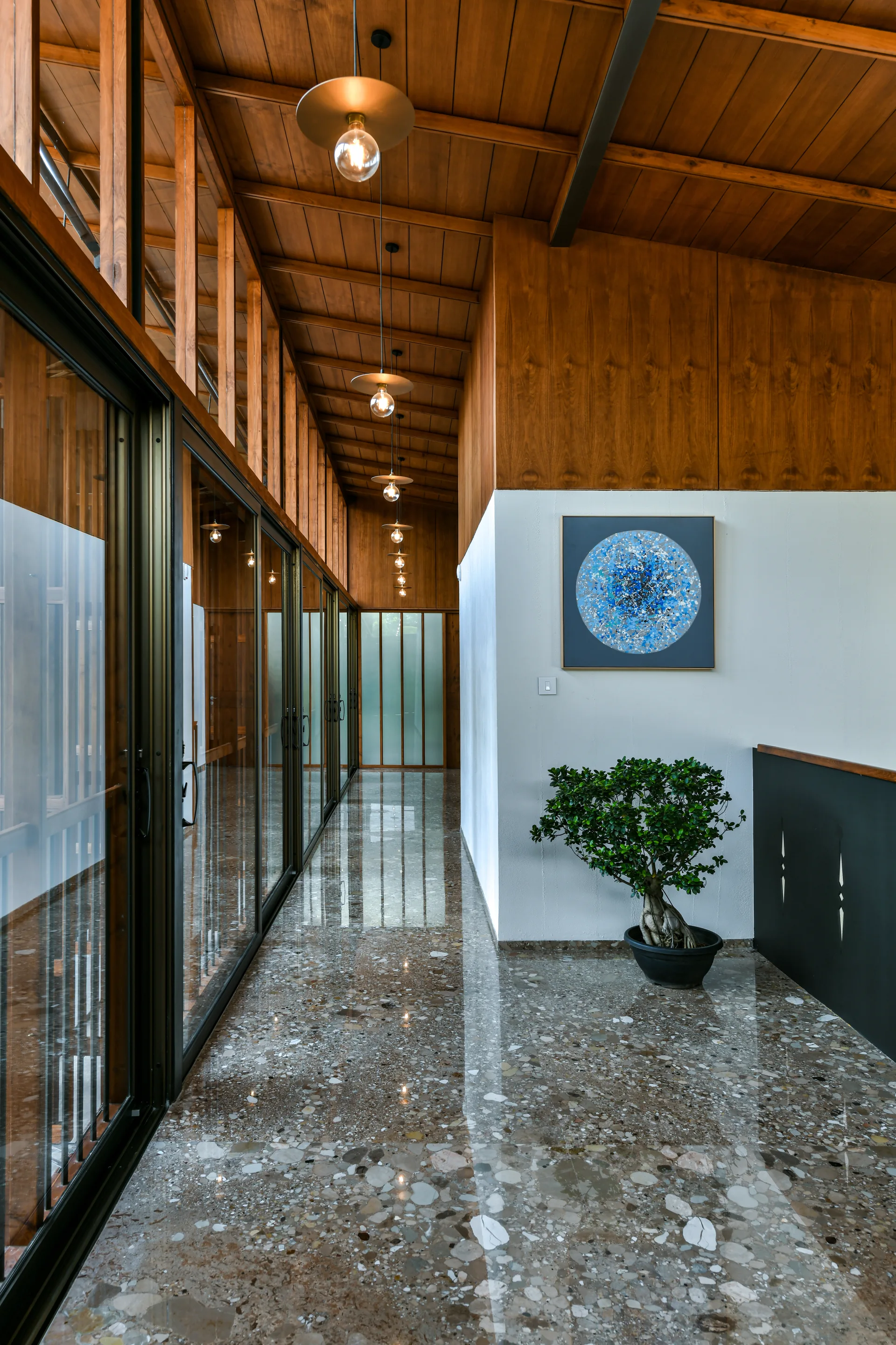 A long hallway with a highly reflective terrazzo floor, a sloped wood ceiling with a row of pendant lights, and sliding glass doors.