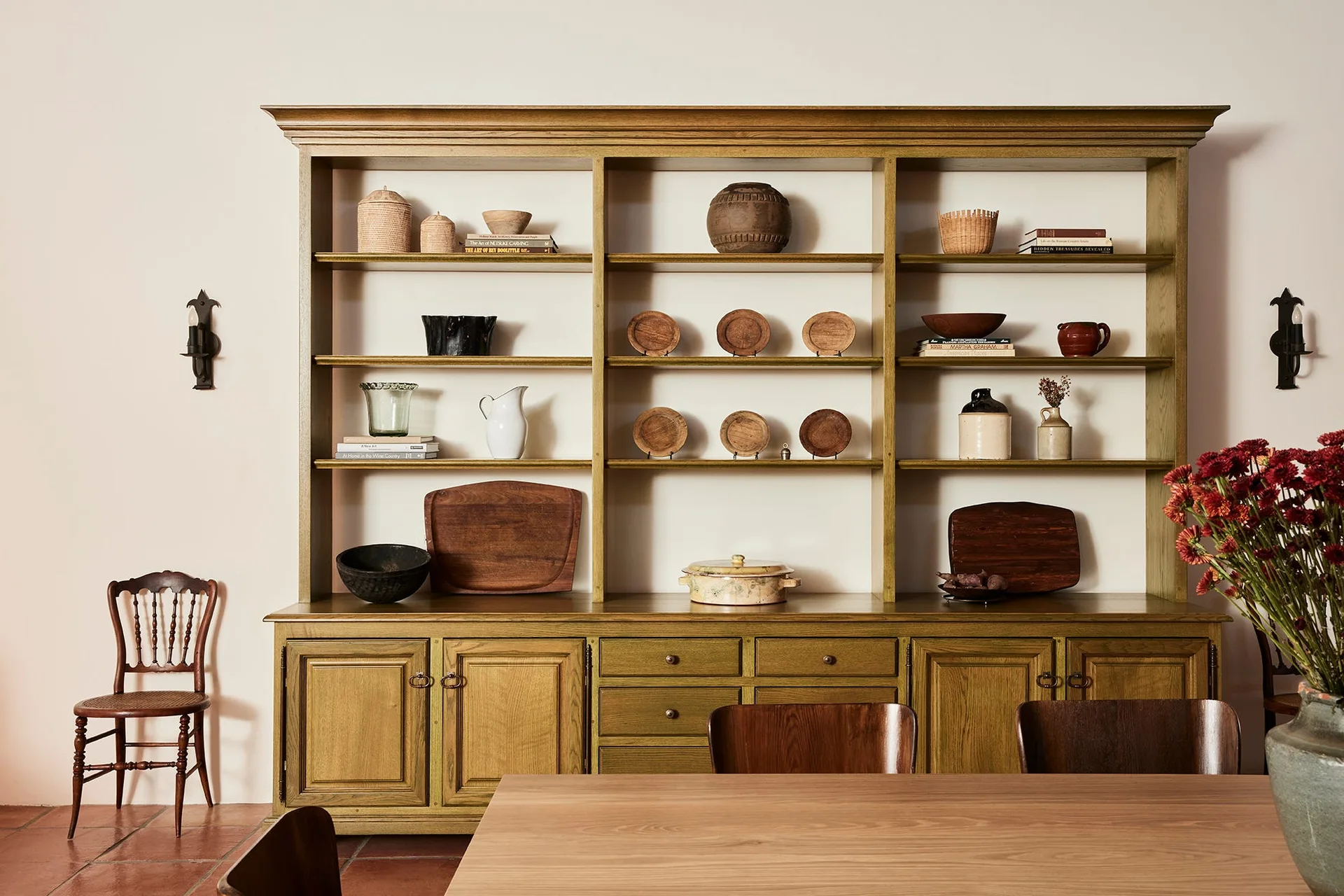 A large olive green wooden hutch with open shelves filled with ceramic plates, wooden cutting boards, and woven baskets sits against a cream wall.