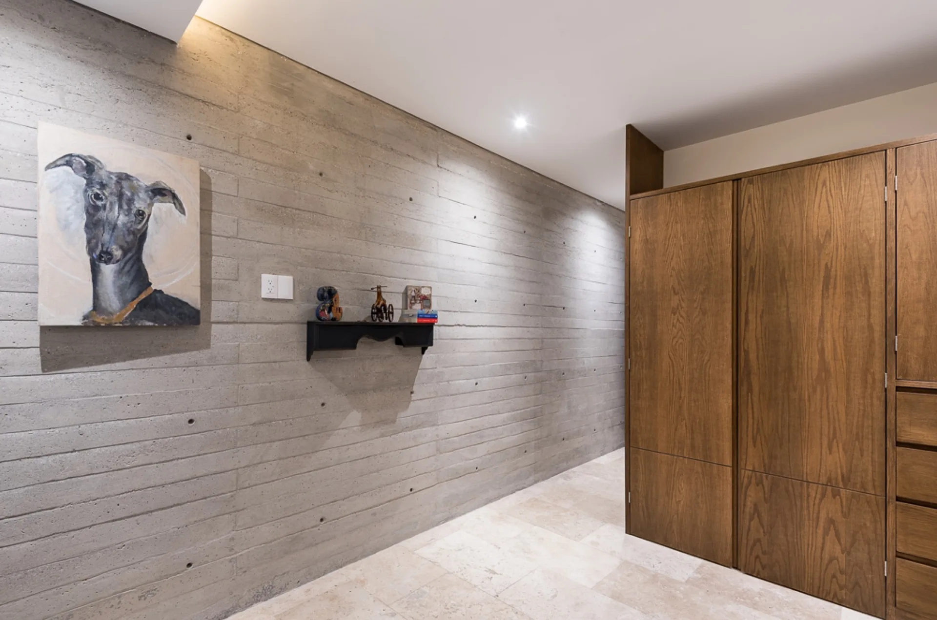 A minimalist hallway featuring a textured board-formed concrete wall, floor-to-ceiling wood cabinetry, and beige travertine tile flooring.