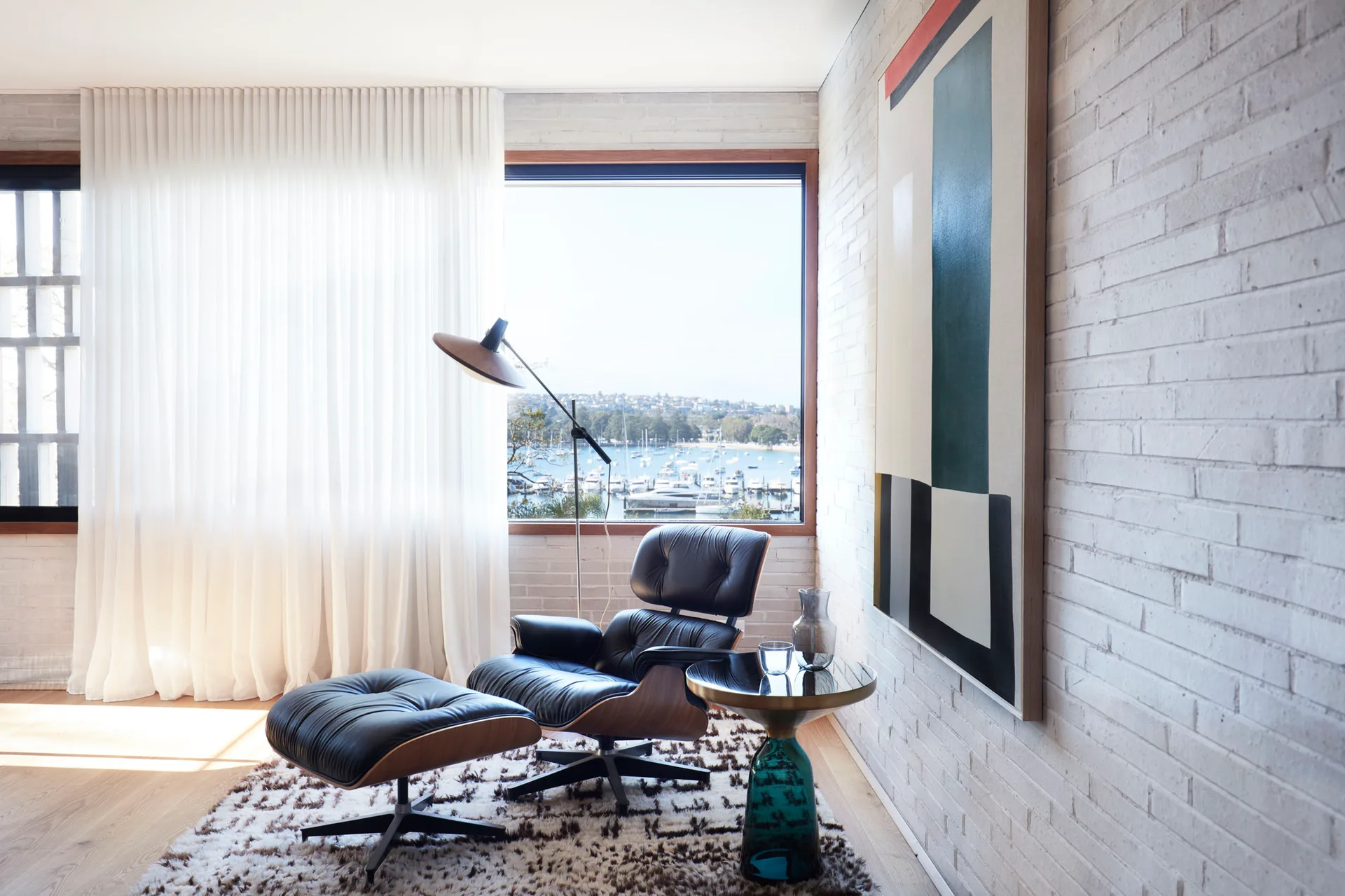 A black leather lounge chair and ottoman sit on a patterned rug next to a white painted brick wall and a window overlooking a harbor.