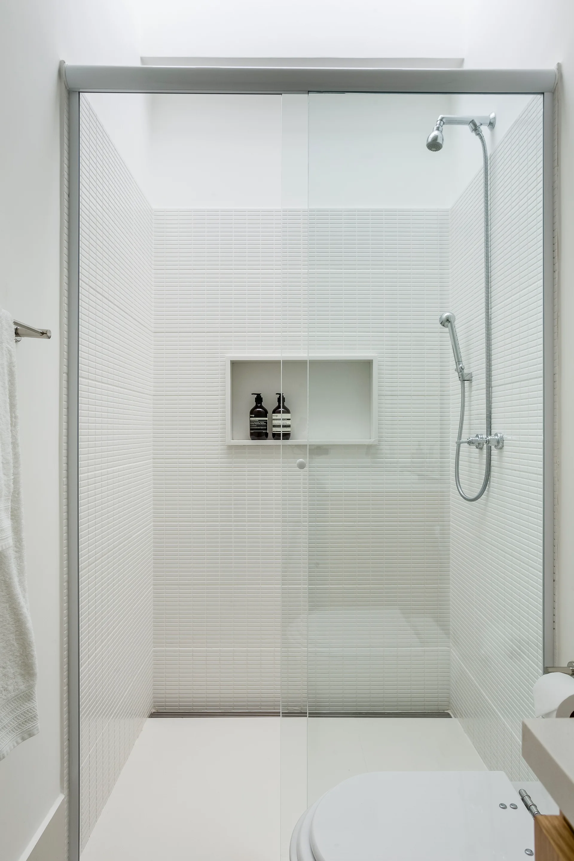 A minimalist white shower features small grid mosaic tiles, a recessed shelf with two bottles, a chrome shower head, and a sliding glass door.