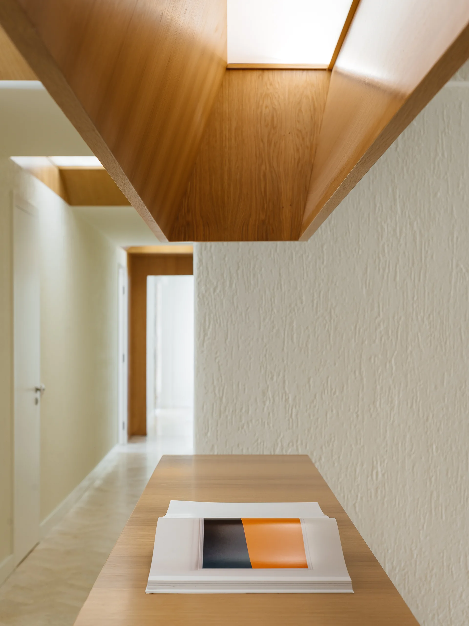 A minimalist hallway features textured plaster walls, a wooden console table in the foreground, and a warm wooden lighting channel recessed in the ceiling.