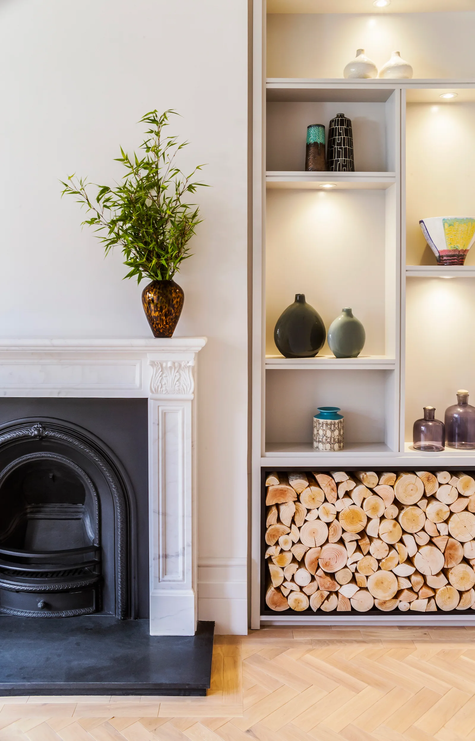 A white marble fireplace stands beside built-in shelving styled with ceramic vases and a bottom compartment filled with neatly stacked firewood.