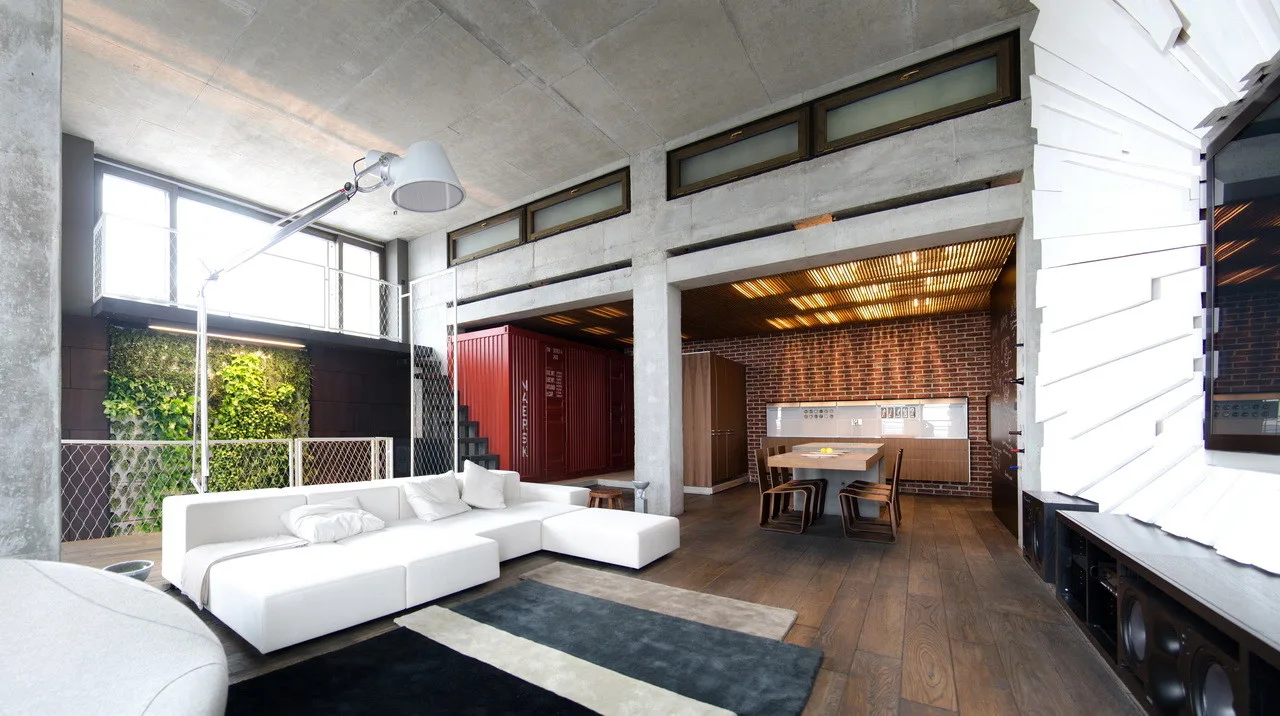 An open-plan industrial loft featuring a white sectional sofa, an indoor living wall, and a red shipping container near a brick-walled kitchen.