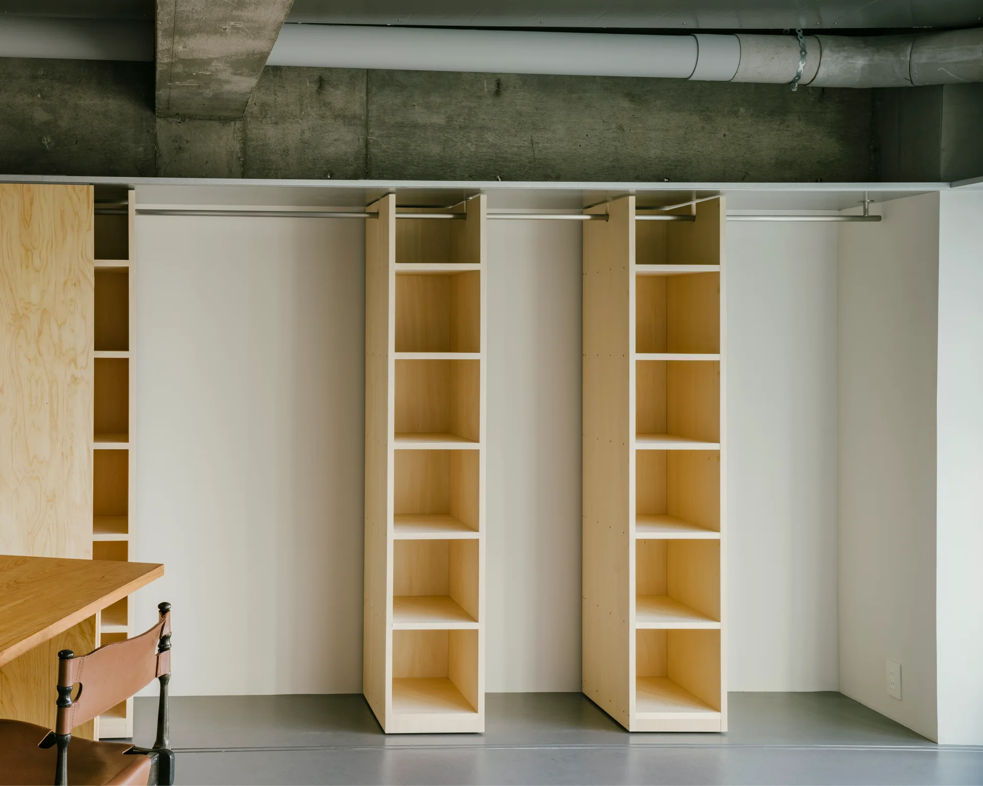 A minimalist open wardrobe featuring light wood shelving towers and a metal clothing rod against a white wall under an exposed concrete ceiling.