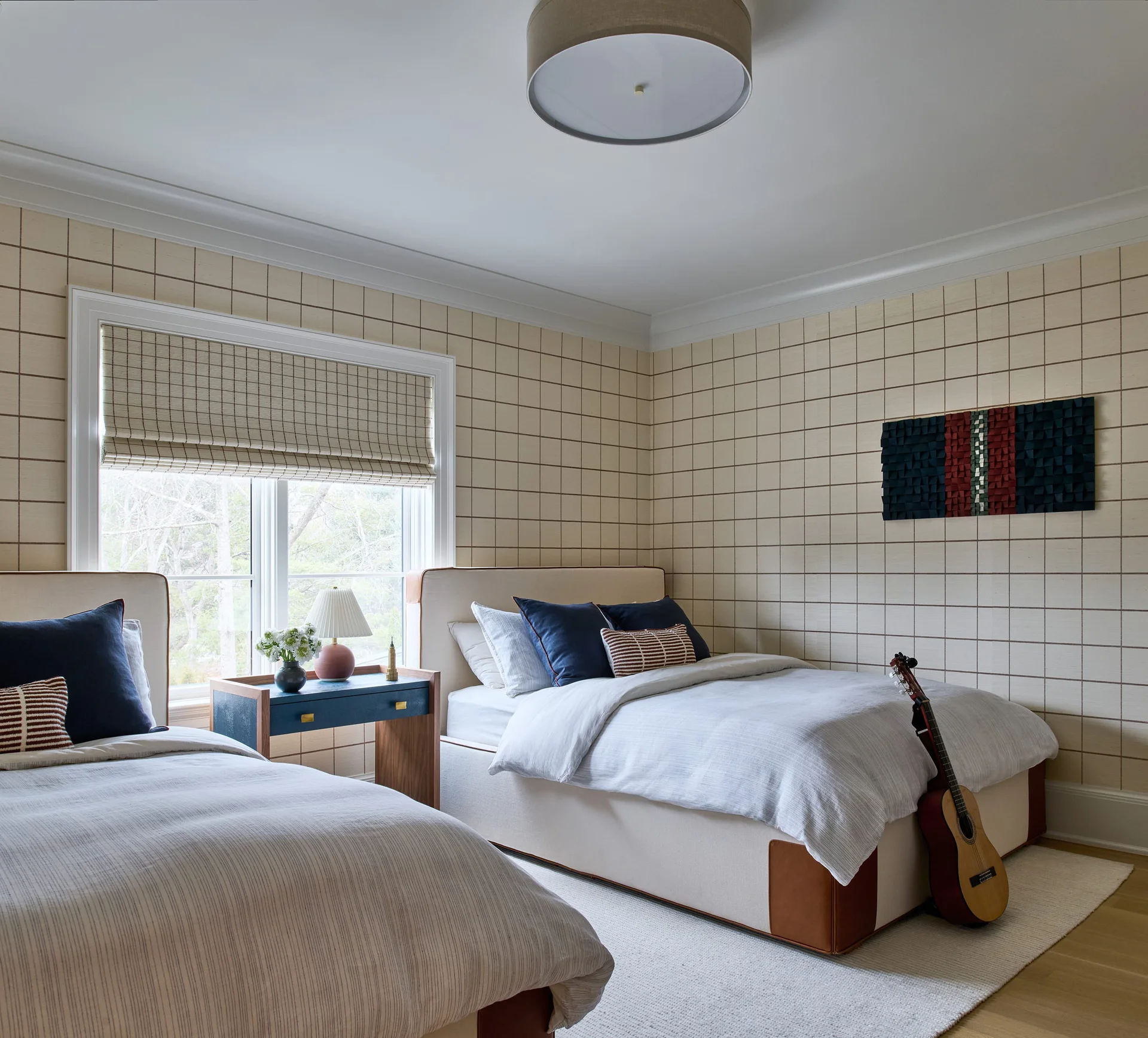 A bright bedroom with two twin beds featuring navy accent pillows, a blue nightstand, and cream grid-patterned wallpaper.