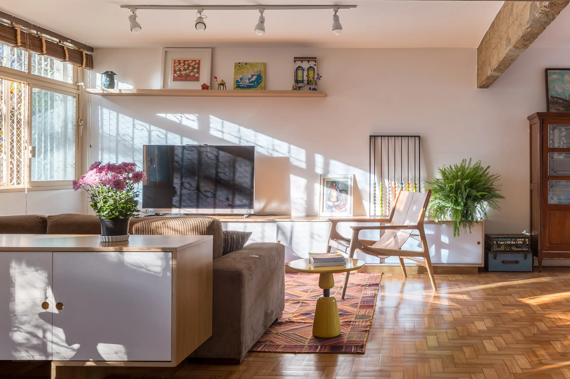 A sunlit living room featuring a brown corduroy sofa, a yellow pedestal side table, and classic herringbone parquet wood flooring.