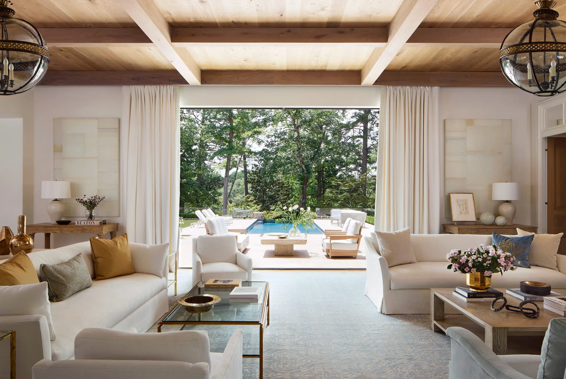 A bright living room with white sofas, exposed wood ceiling beams, a patterned blue rug, and large sliding doors that open to an outdoor pool terrace.