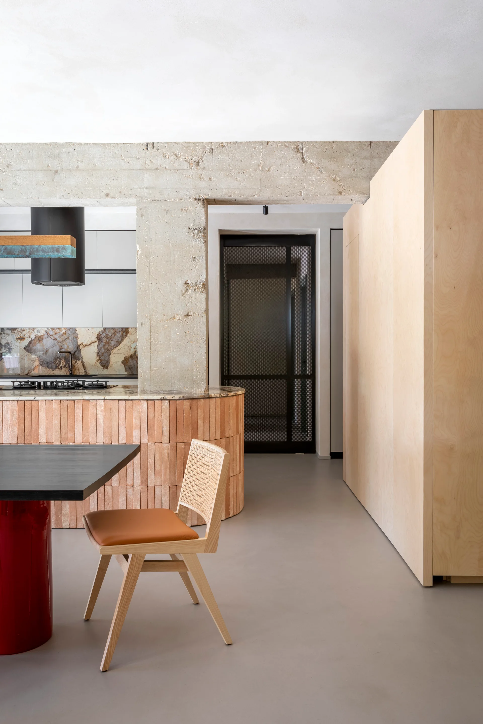 A modern kitchen showing a curved island with vertical terracotta tiles, a light wood storage cabinet, and a cane-backed dining chair.