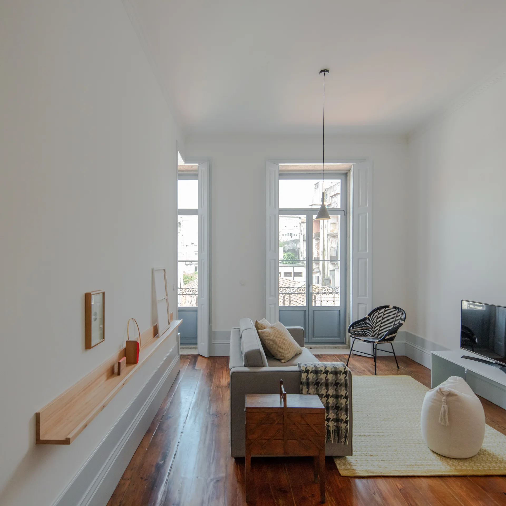 A minimalist white living room features a gray sofa, polished wood floors, tall balcony windows, and a long wooden display ledge on the wall.