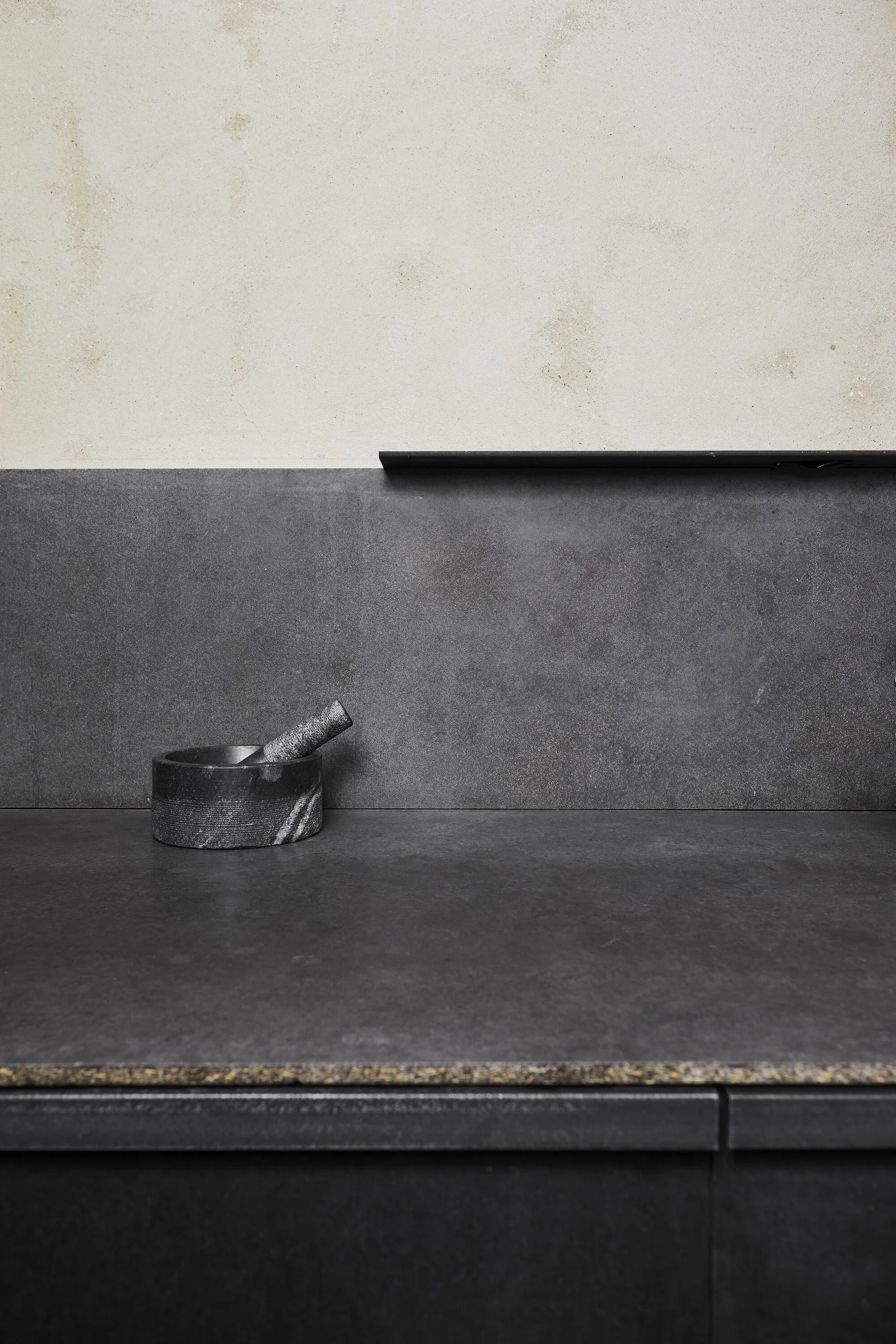 A close-up of a charcoal stone kitchen countertop and backsplash with a mortar and pestle below a light plaster wall and thin black shelf.