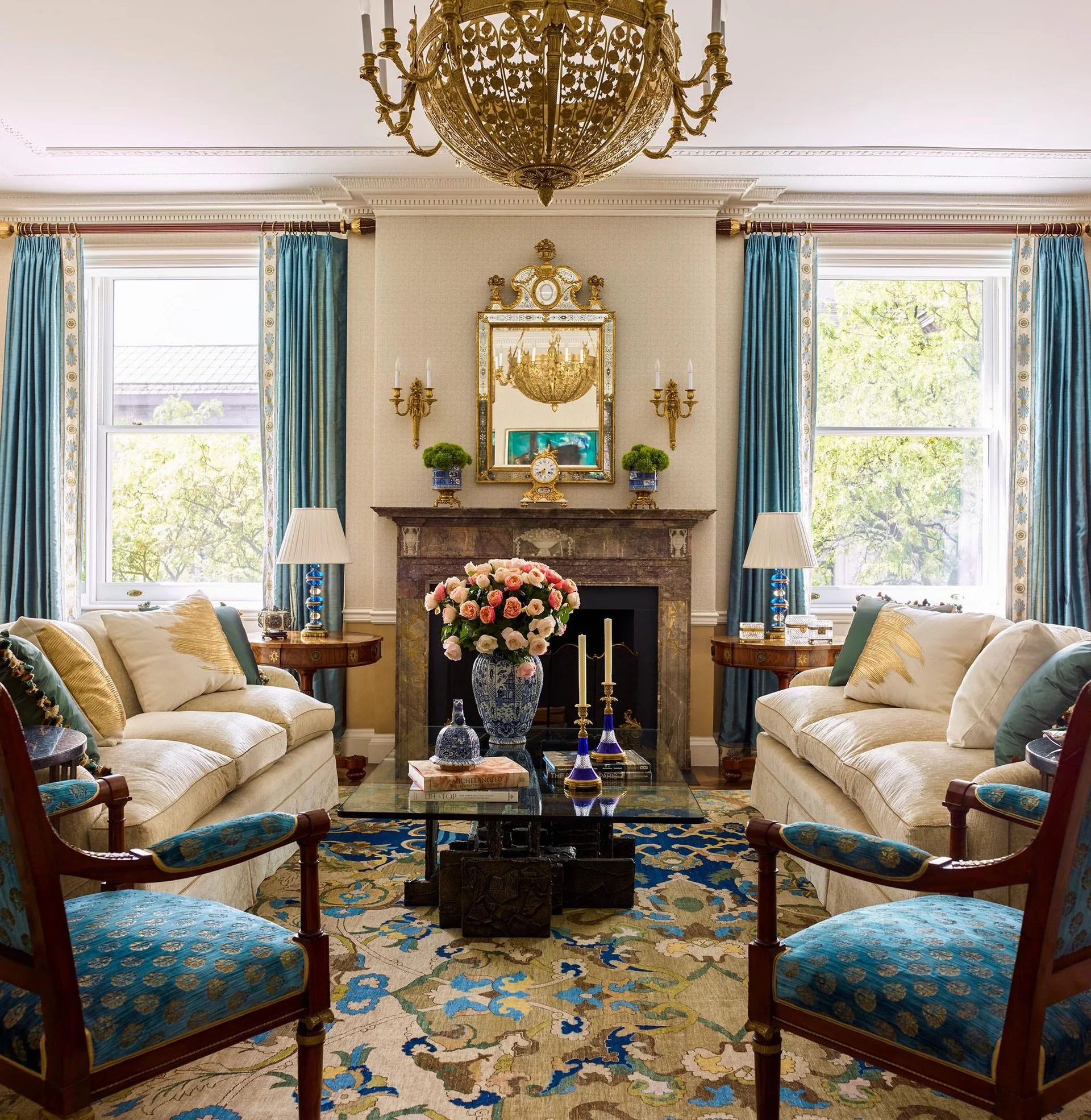 A symmetrical traditional living room featuring two cream sofas, a central fireplace with an ornate gold mirror, blue drapes, and a patterned rug.