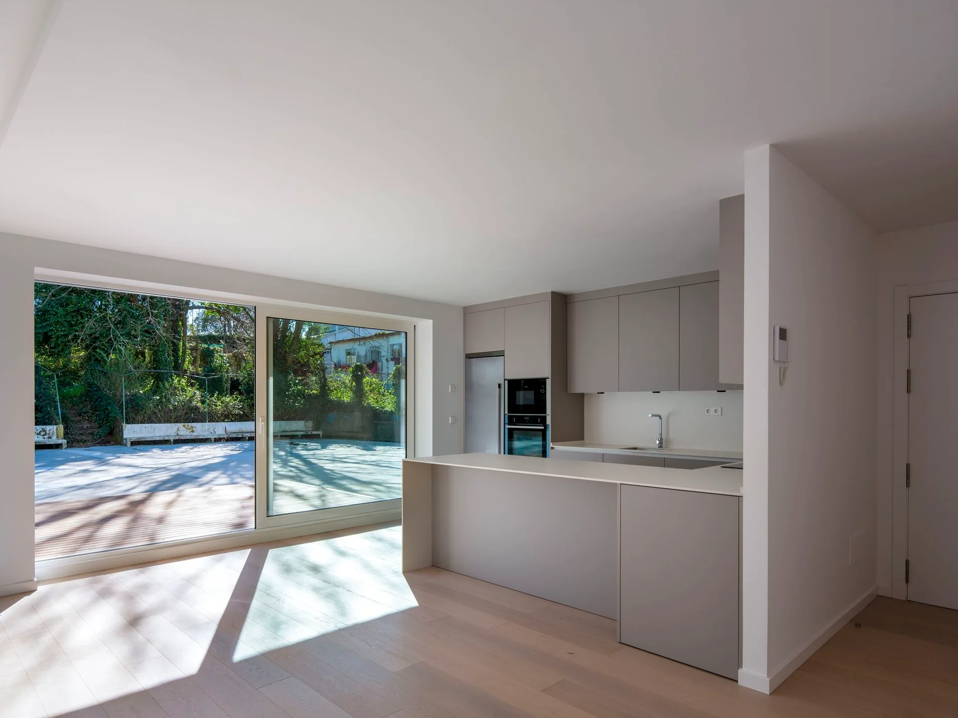A minimalist kitchen features matte gray cabinetry, a matching island, light wood flooring, and a large sliding glass door leading outside.
