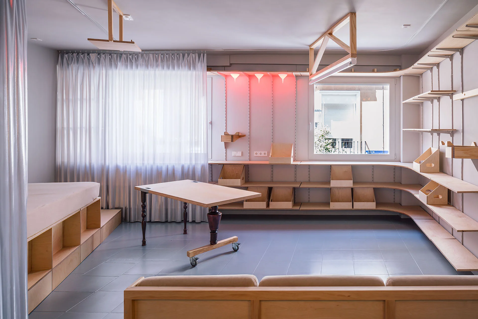 A multi-functional studio room features a modular plywood shelving system, a mobile wooden table with wheels, and gray floor tiles.
