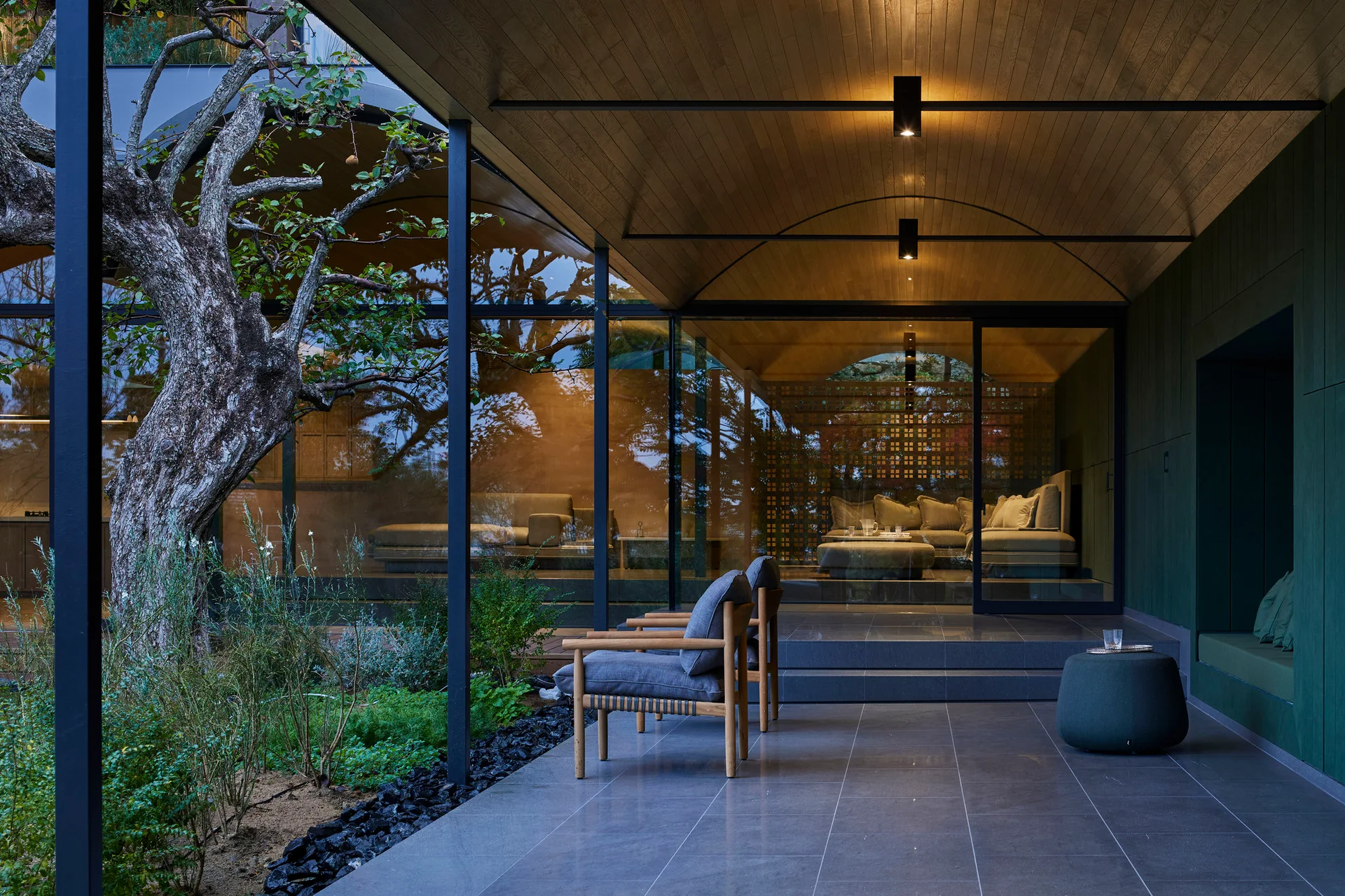Two wooden armchairs sit on a grey stone tile patio under a curved wood ceiling next to a glass wall showing a living room inside.
