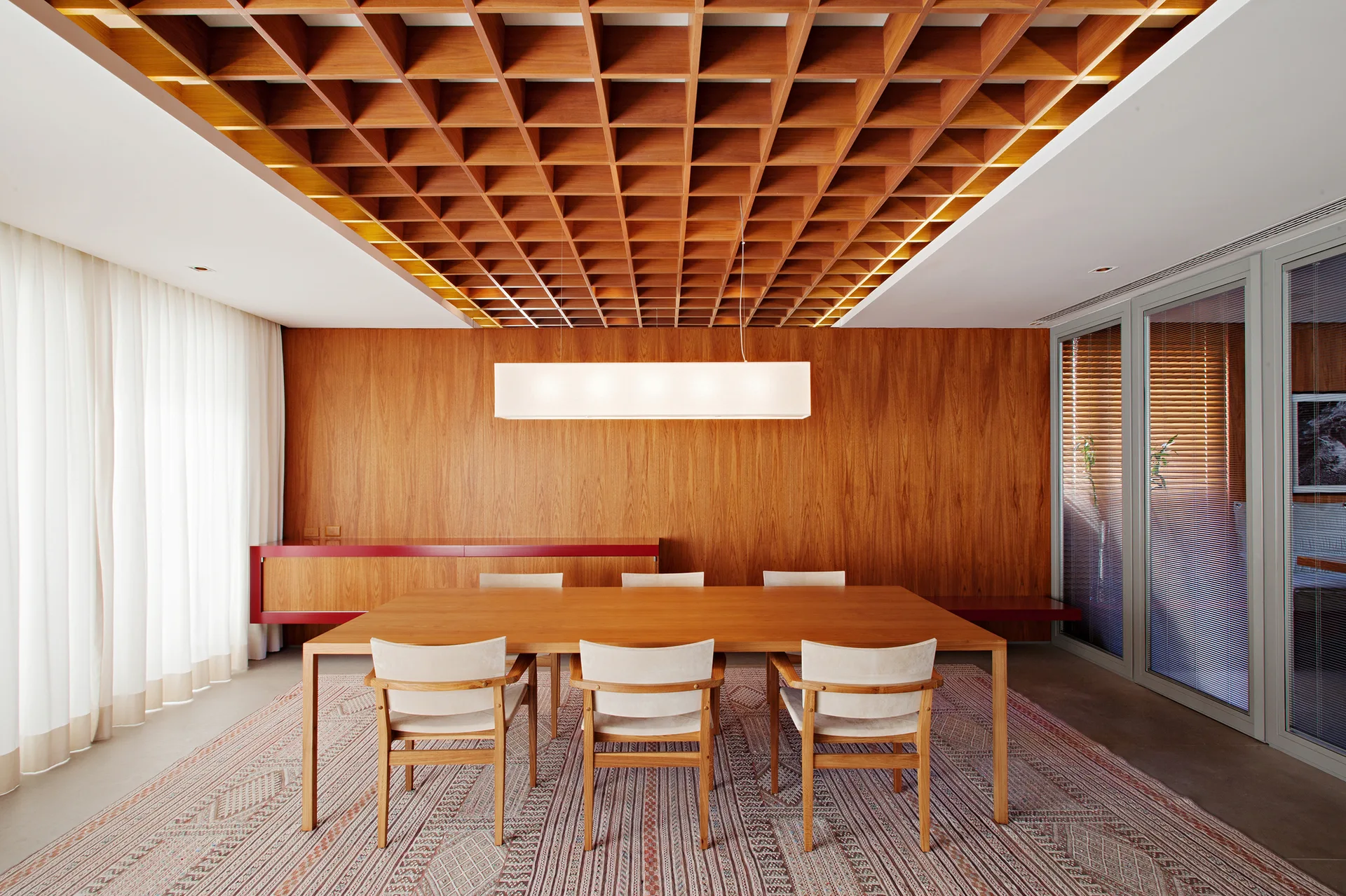 A dining room features a large wooden table on a patterned rug beneath a geometric, coffered wood ceiling and a rectangular pendant light.