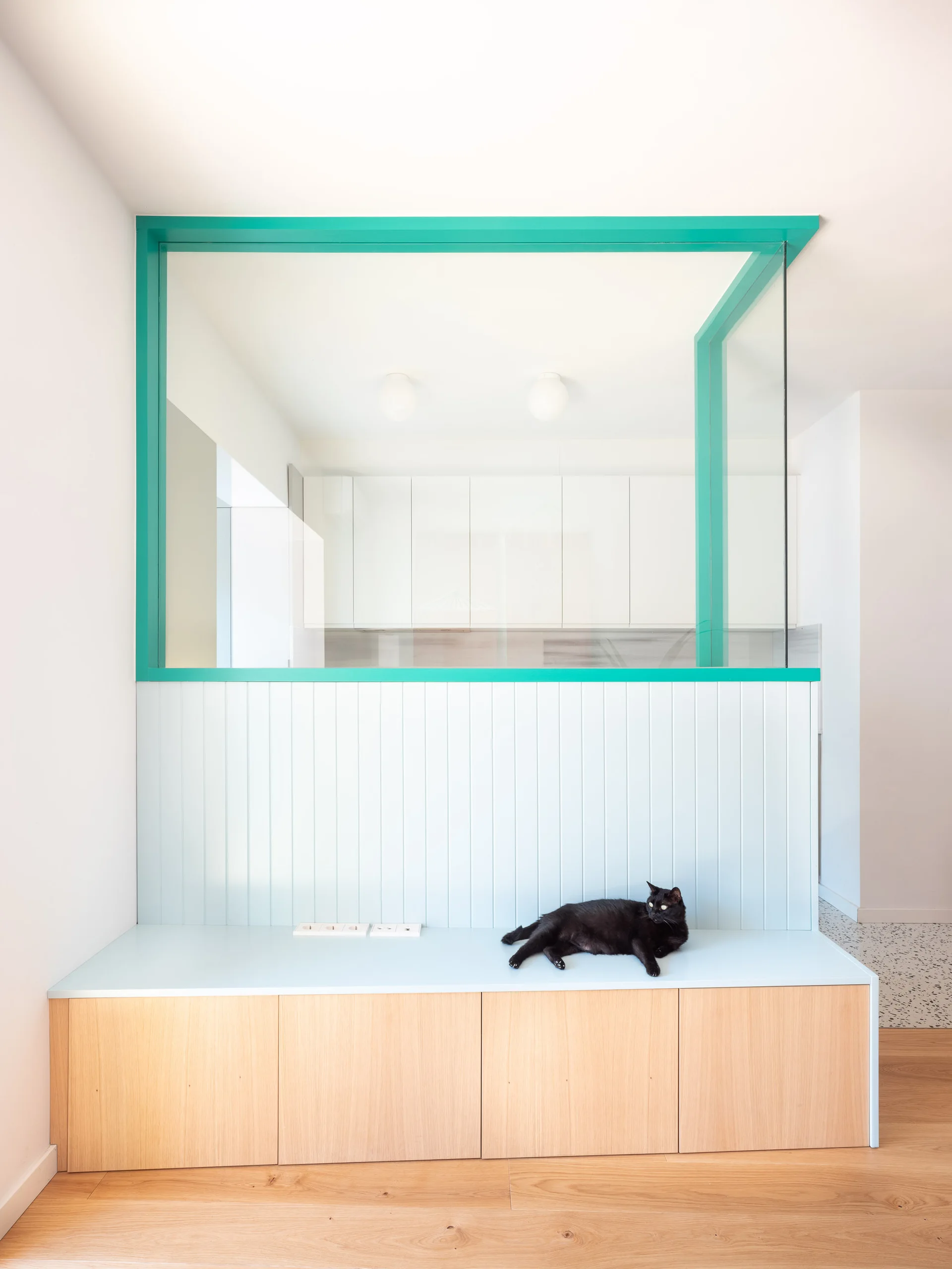 A modern light wood storage bench is topped with pale blue paneling and a teal-framed glass partition looking into a white kitchen.