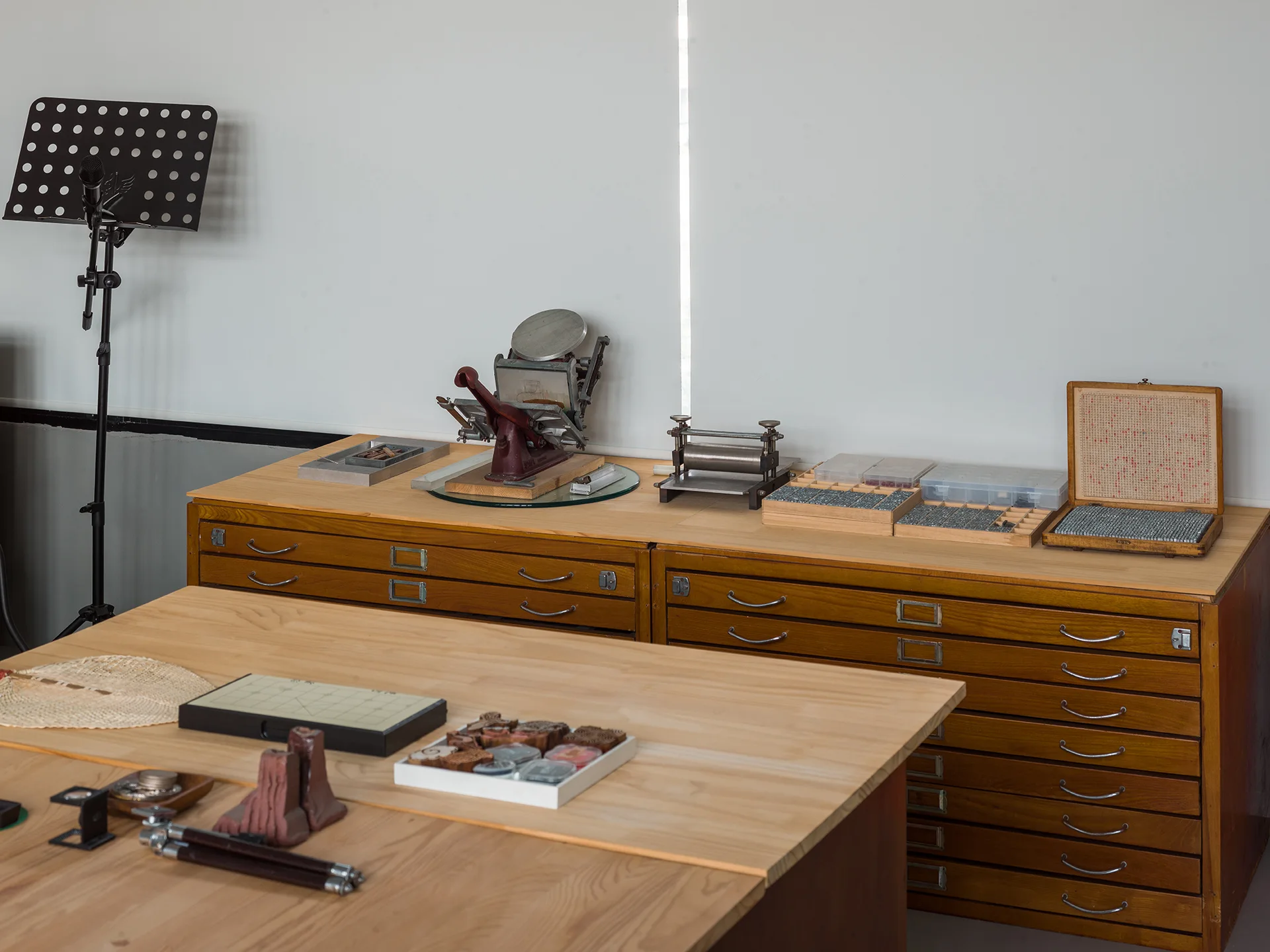 A wooden storage cabinet with shallow drawers and a work table with craft tools sit against a white window shade.